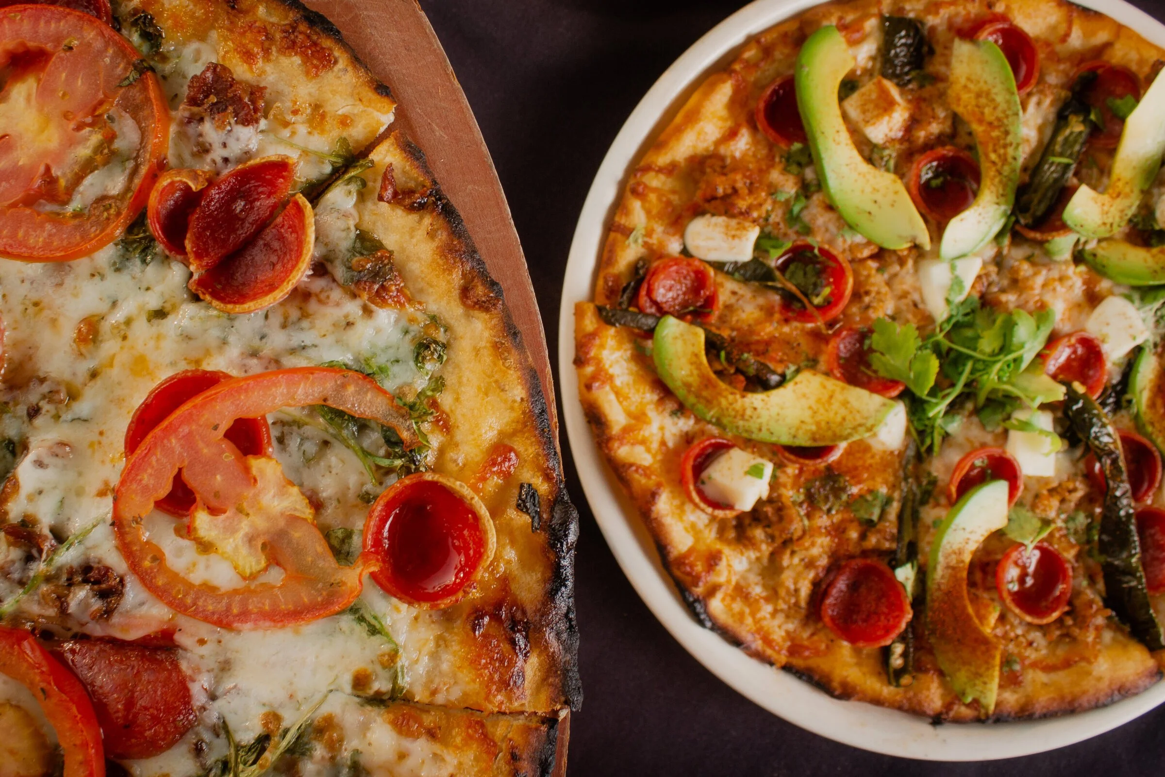 Two pizzas with various toppings, including tomatoes, green peppers, and cheese, on a dark surface.