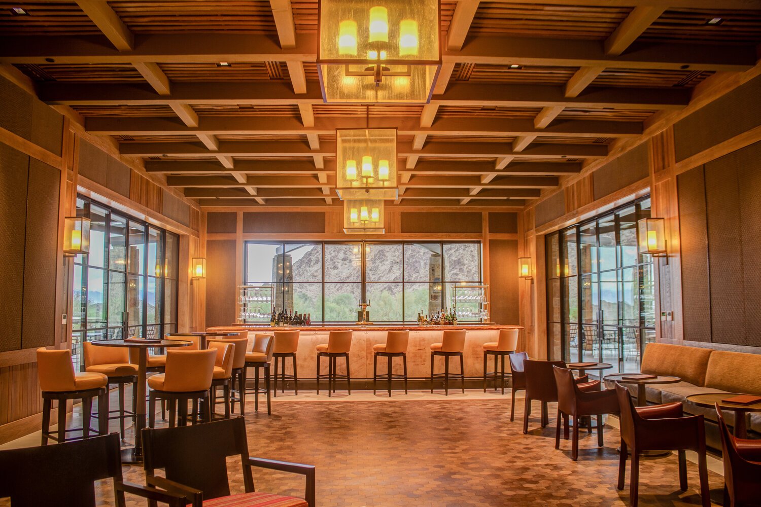 Interior of a modern bar and lounge with high ceiling, wooden beams, and warm lighting. Bar counter with stools, large windows with views of mountains, and seating areas with chairs and tables.