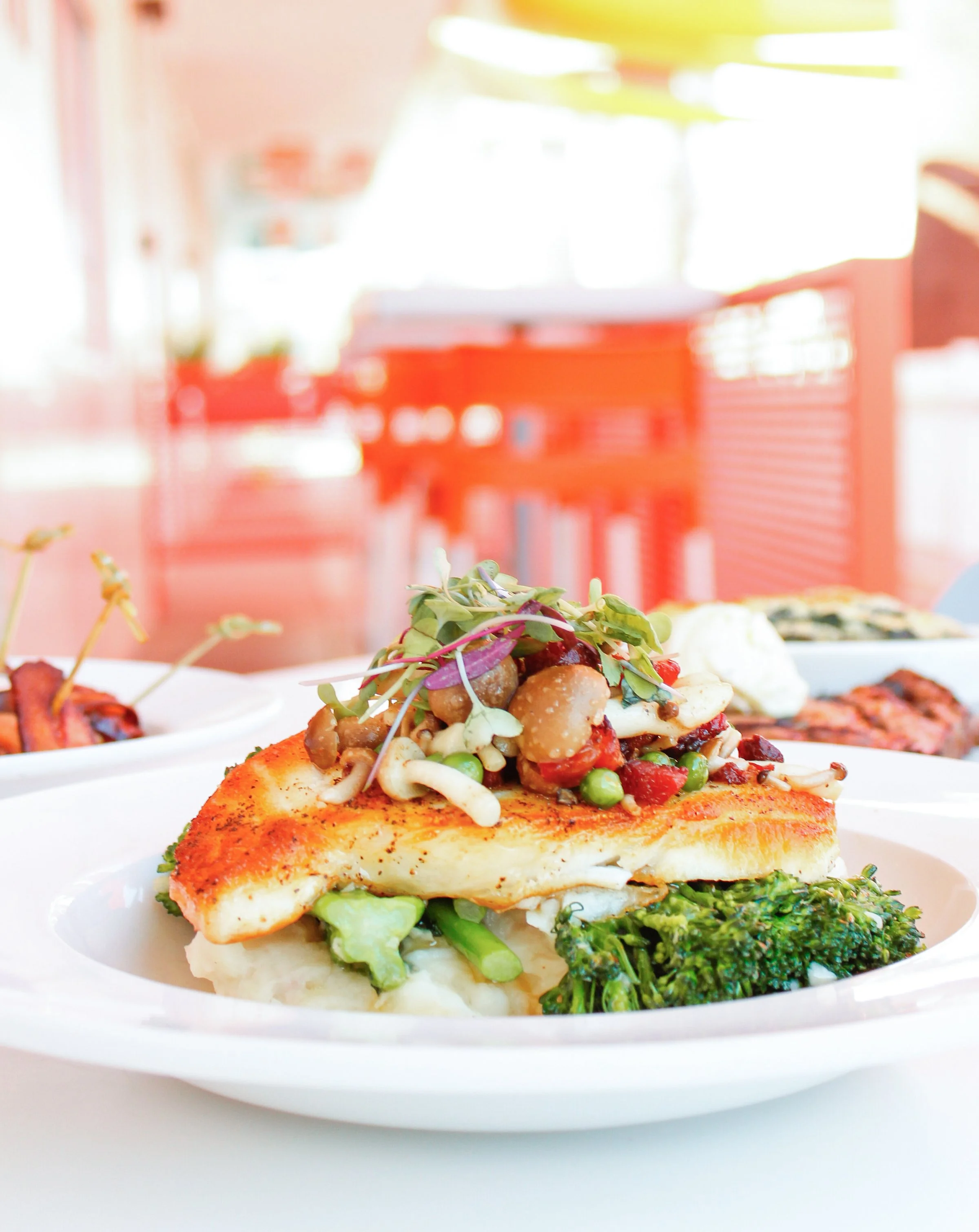 A plated entree with cooked fish topped with a colorful vegetable medley, served with broccoli and mashed potatoes, on a white plate in a brightly lit restaurant or cafe setting.