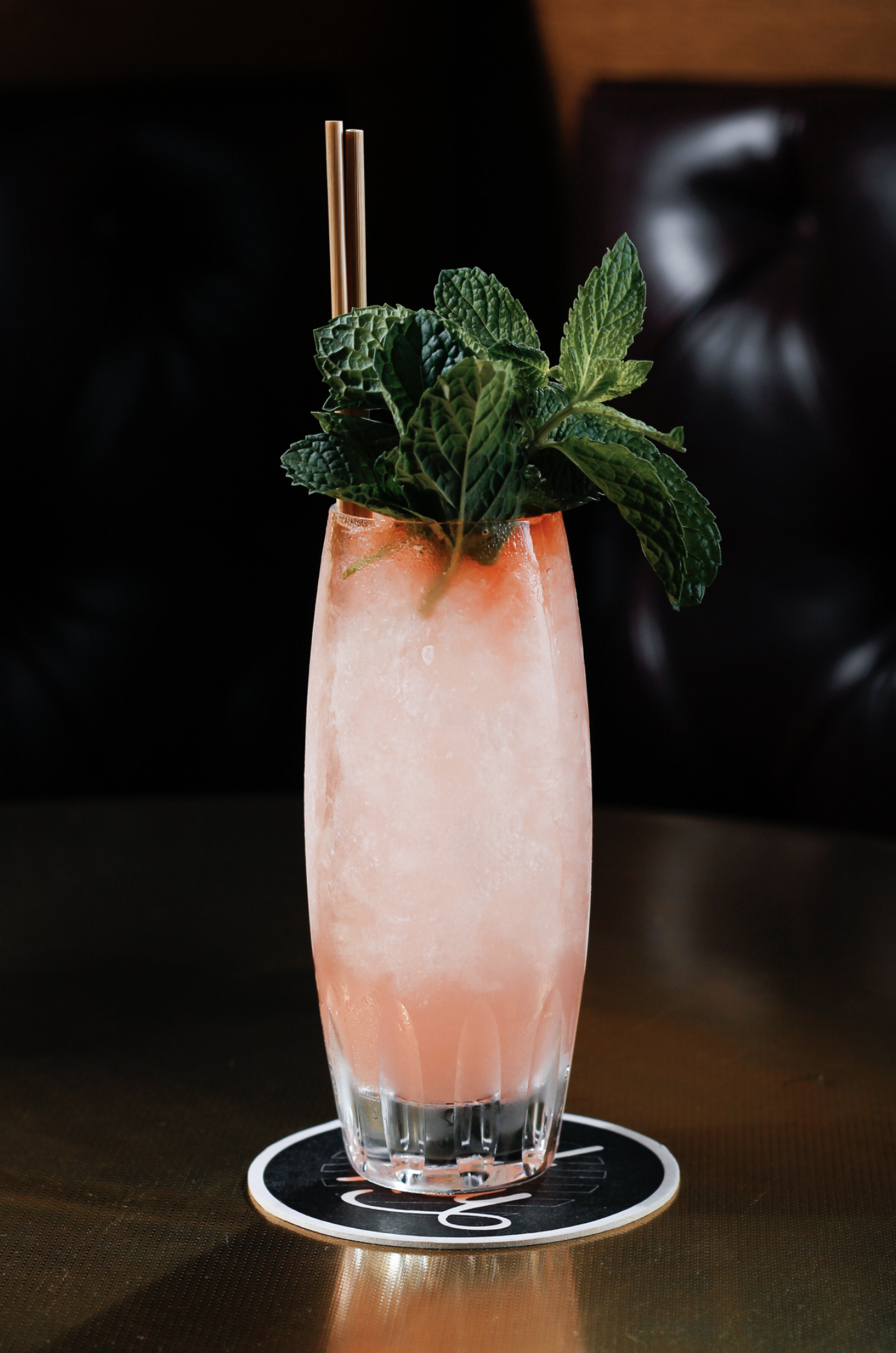 A tall glass of pink cocktail garnished with fresh mint leaves and a bamboo straw, placed on a black and white coaster on a wooden table.
