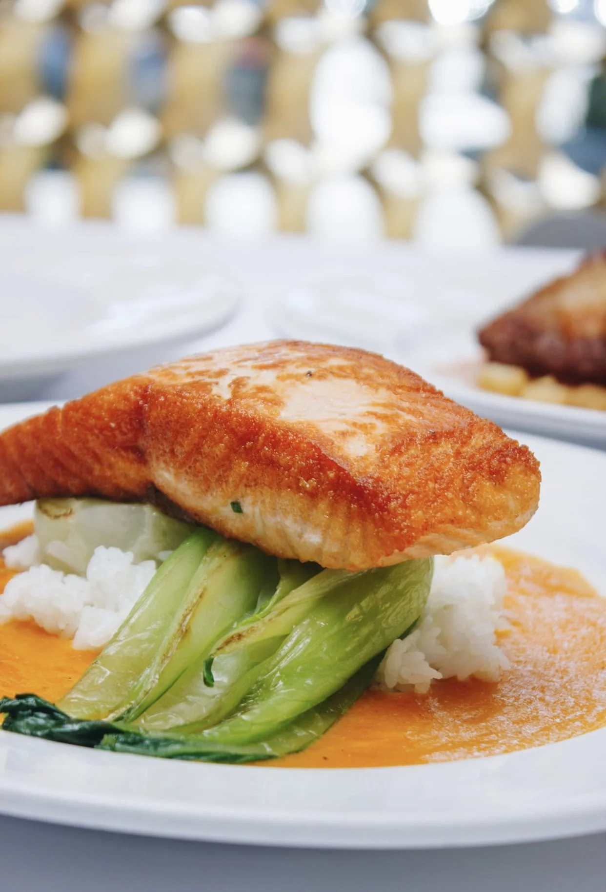A plate of fried fish served with vegetables and sauce, with another plate of food in the background.