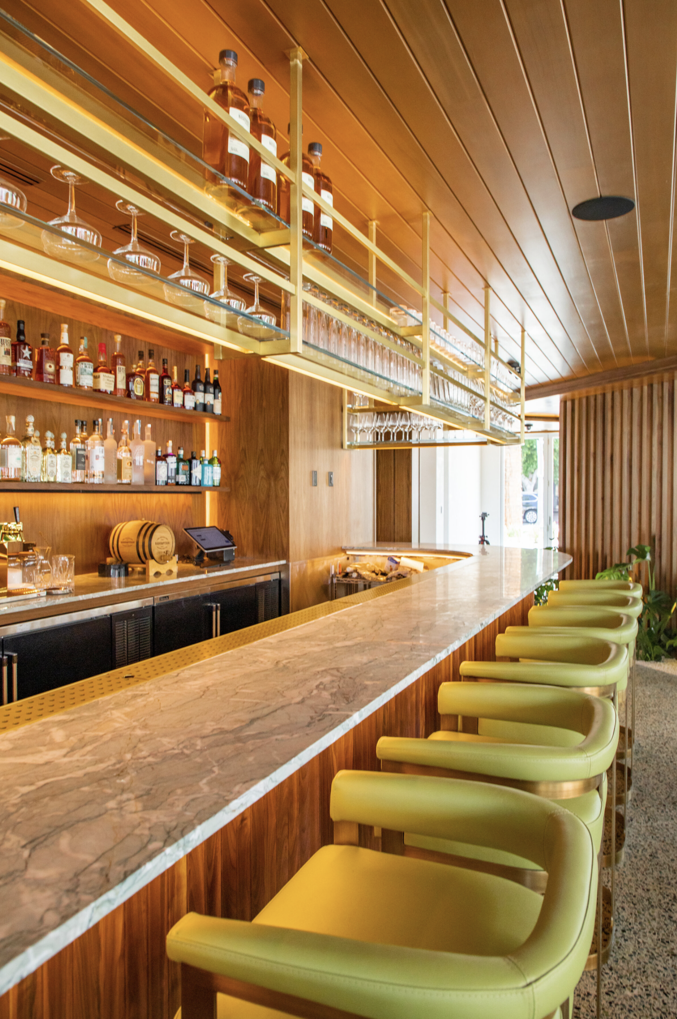 A modern bar with a marble countertop, wooden shelves stocked with liquor bottles, and green bar stools.