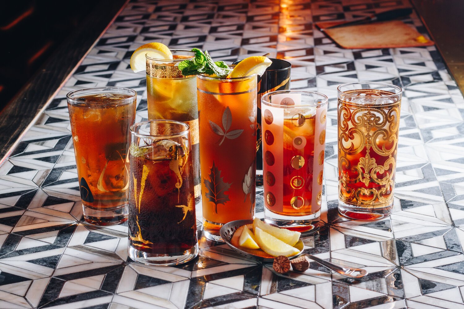 Six glasses of iced tea with lemon and mint, on a patterned table, with lemon wedges and a spoon in front.