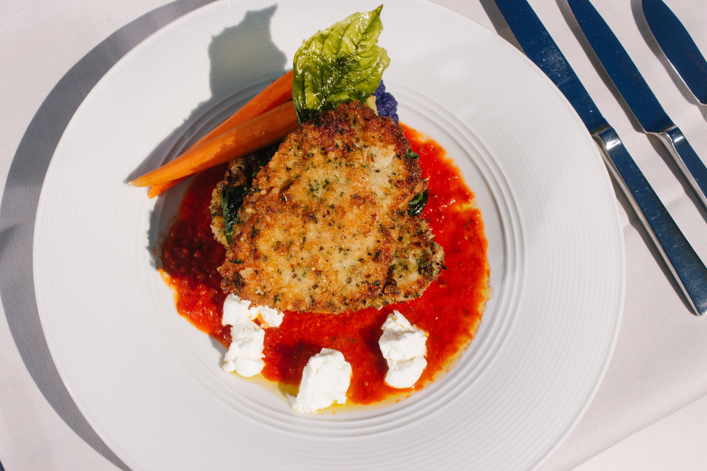Fried breaded fish fillet on a white plate with tomato sauce, cheese, carrots, lettuce, and purple cauliflower, with a fork and knife beside the plate.