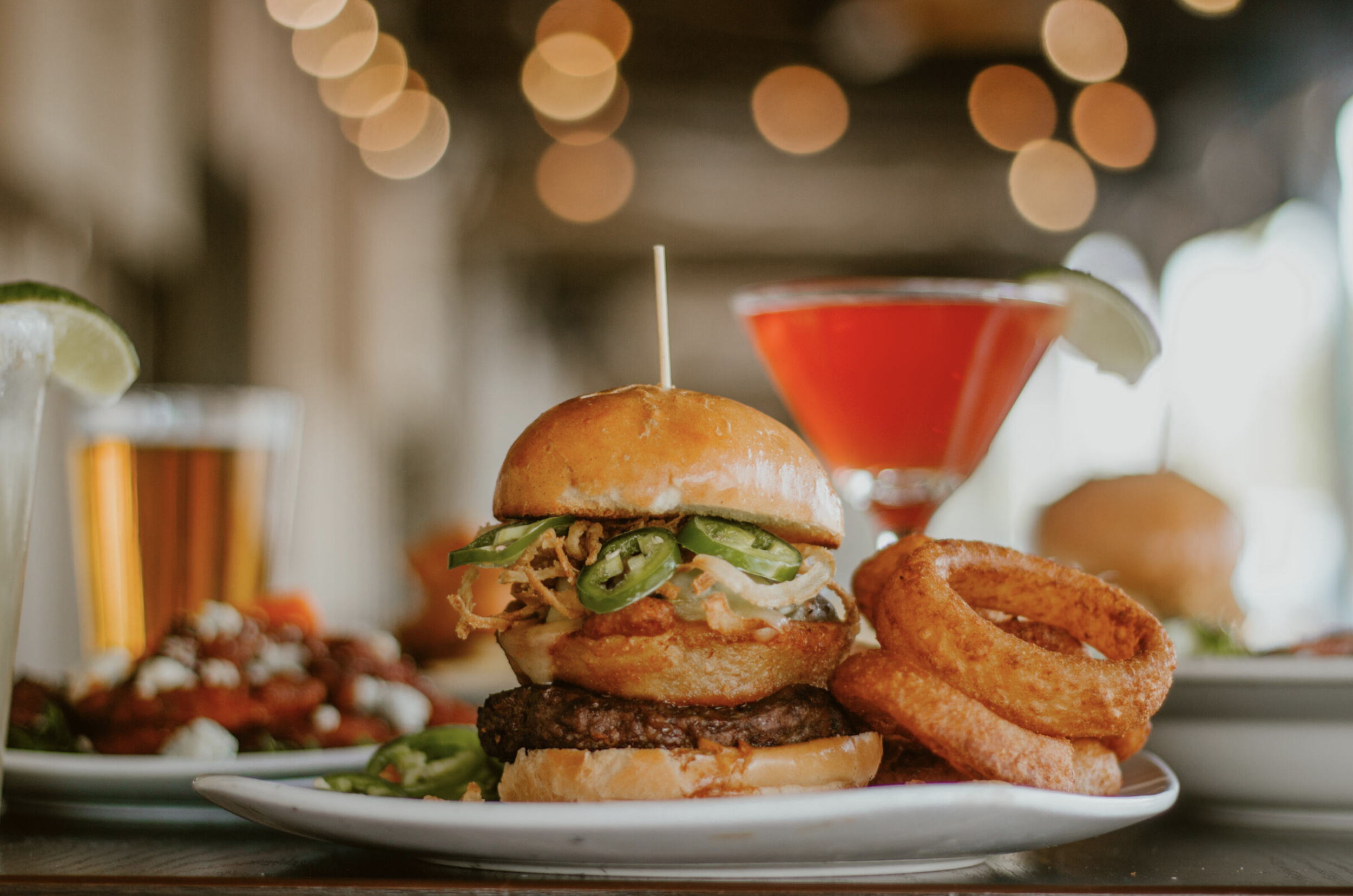 A plate with a hamburger topped with jalapenos, a fried onion ring, and a beef patty, accompanied by a side of onion rings. In the background, there is a cocktail with a lime wedge, and a blurred cocktail with a yellow peel garnish, all on a table wi
