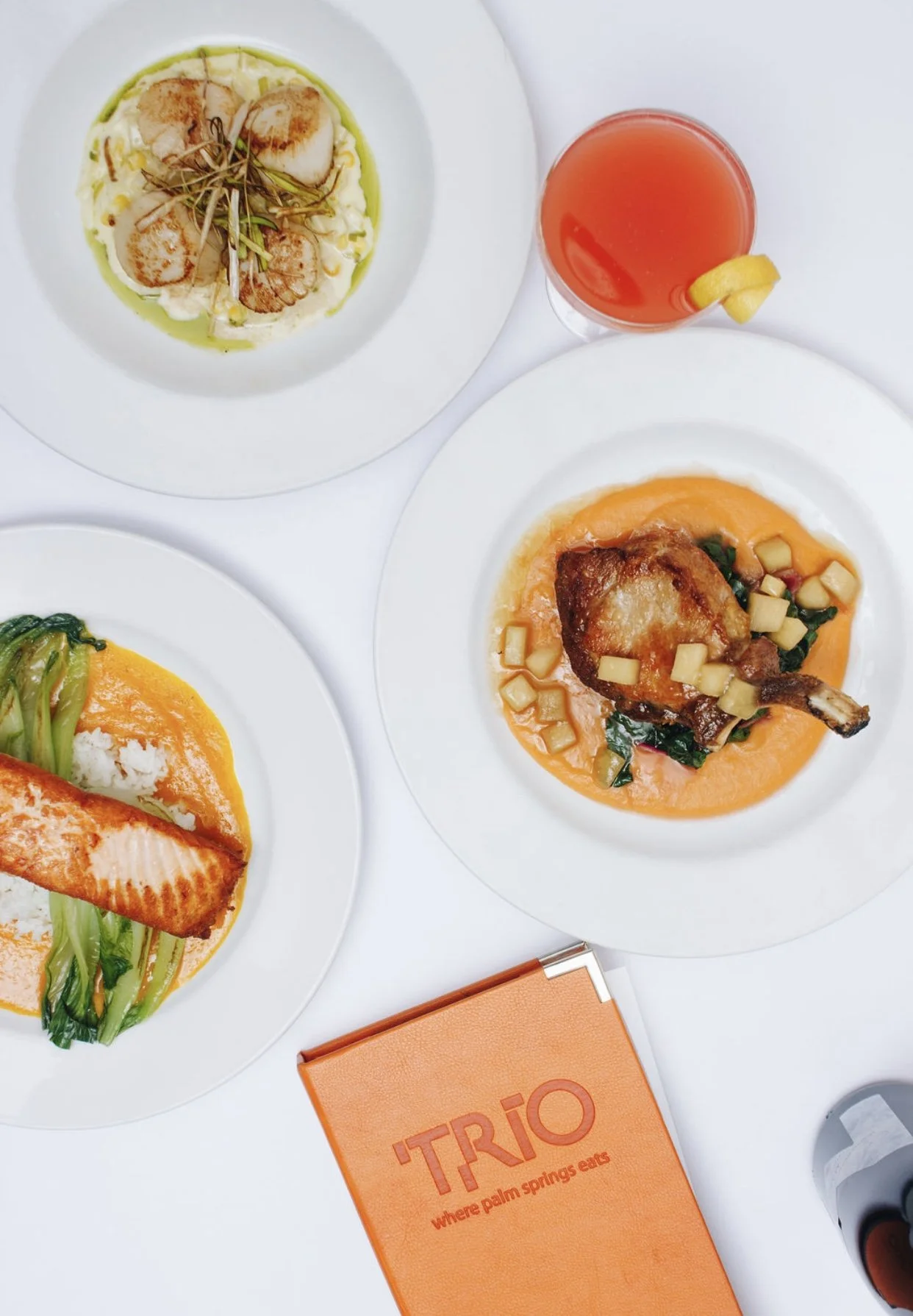 Three plates of food with a drink and a bright orange menu on a white table. The top plate has scallops garnished with herbs, the right plate has a piece of crispy chicken on a bed of orange sauce and diced vegetables, and the left plate has grilled 