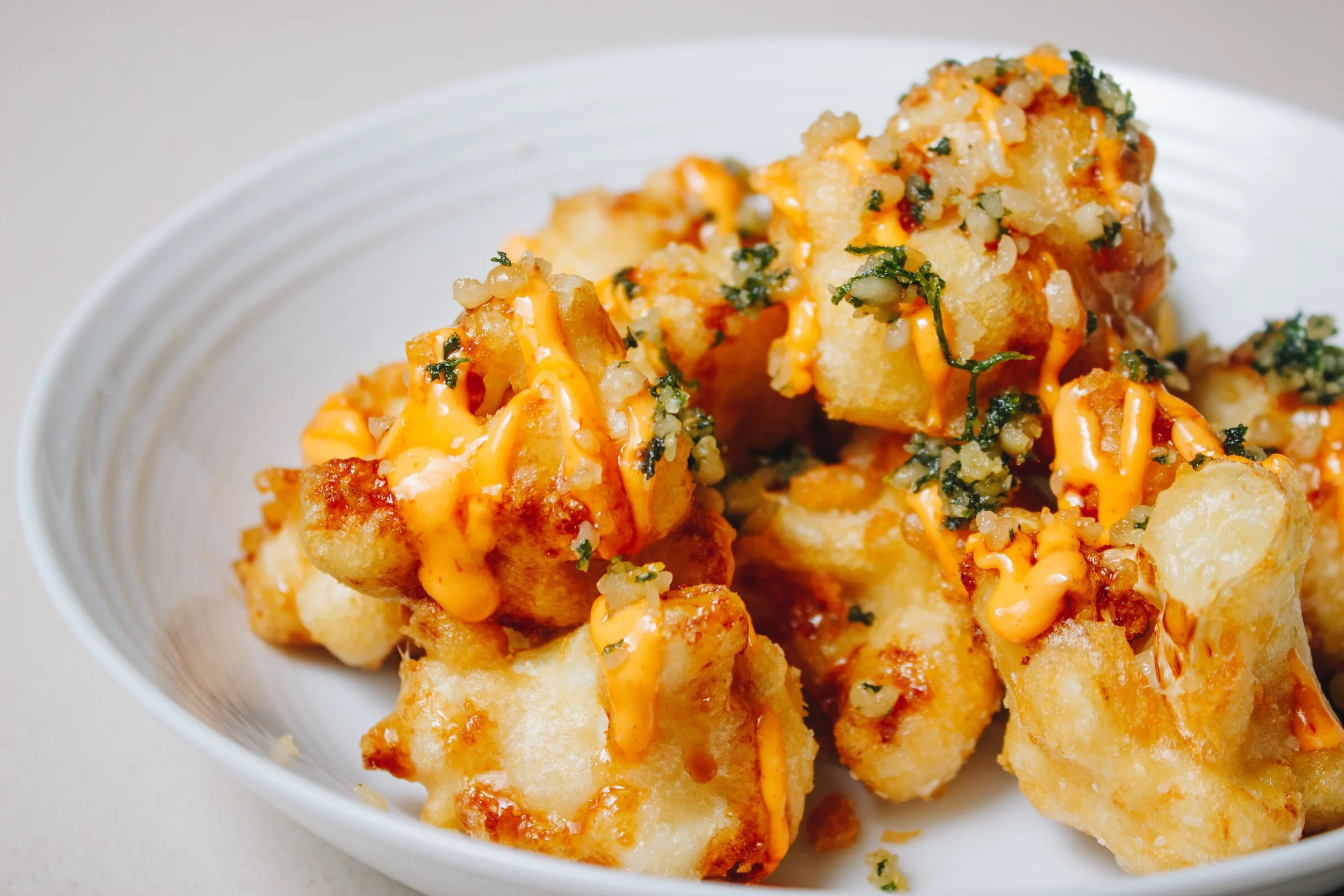 Fried potato chunks topped with spicy sauce, chopped herbs, and cheese in a white bowl.