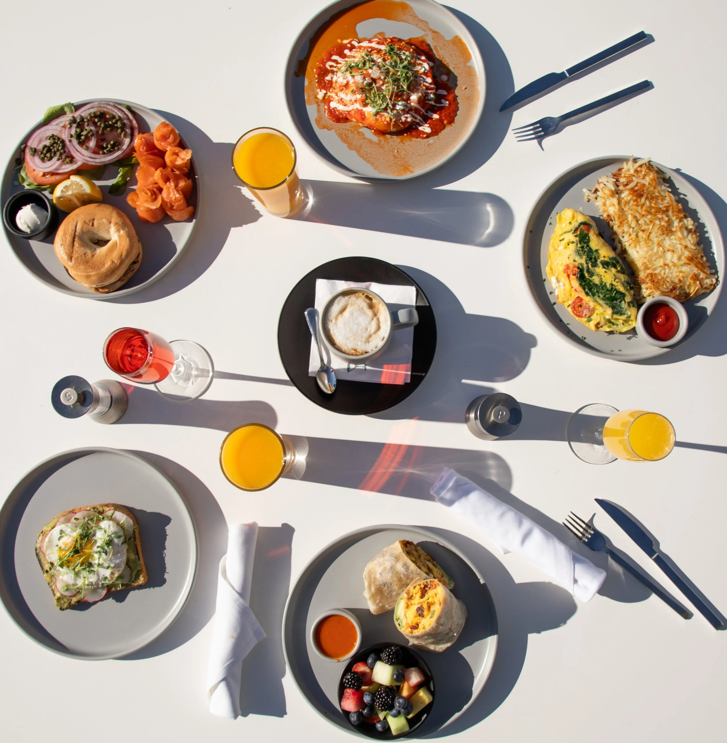 Overhead view of a breakfast table with various dishes, drinks, and utensils. Contains plates of eggs, hash browns, bagels, smoked salmon, fruit salad, and pastries, along with glasses of orange juice, a glass of pink drink, and a coffee cup, set on 