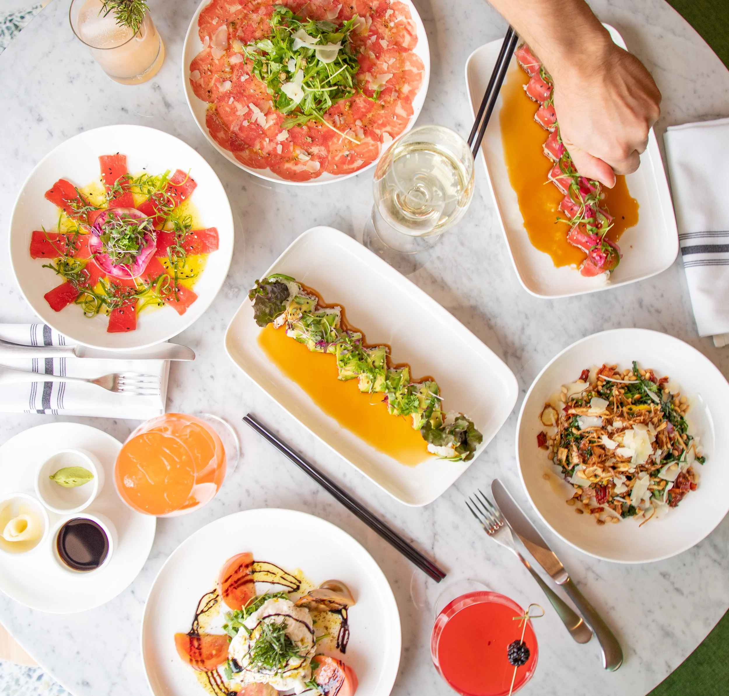 A variety of Asian dishes served on a white marble table, including sashimi, sushi rolls, salad, and drinks.