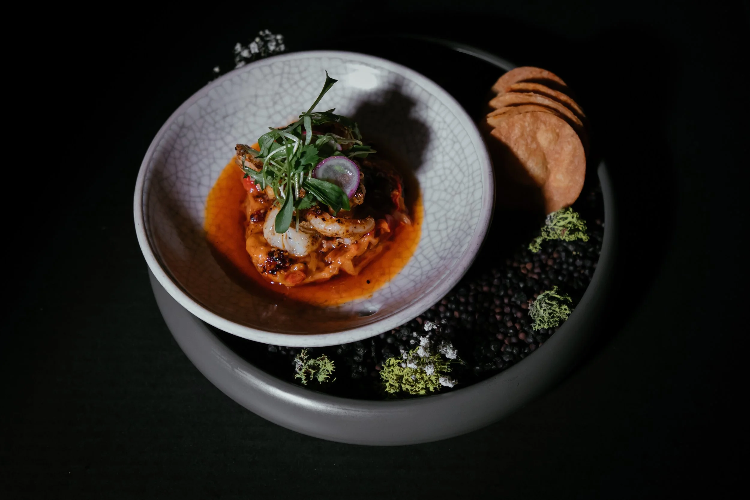 A gourmet dish of grilled seafood topped with microgreens, served in a textured bowl with a red sauce. It is accompanied by crispy round chips and a side of black lentils garnished with greens on a modern black plate.
