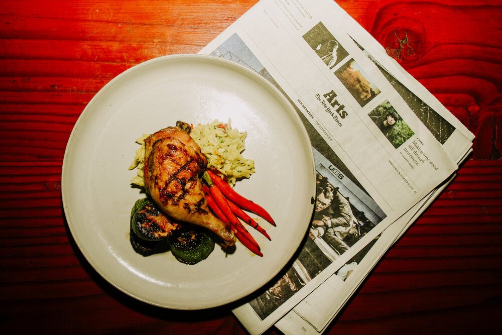 A plate with grilled chicken leg, mashed potatoes, red chili peppers, and grilled zucchini slices next to a newspaper on a wooden table.