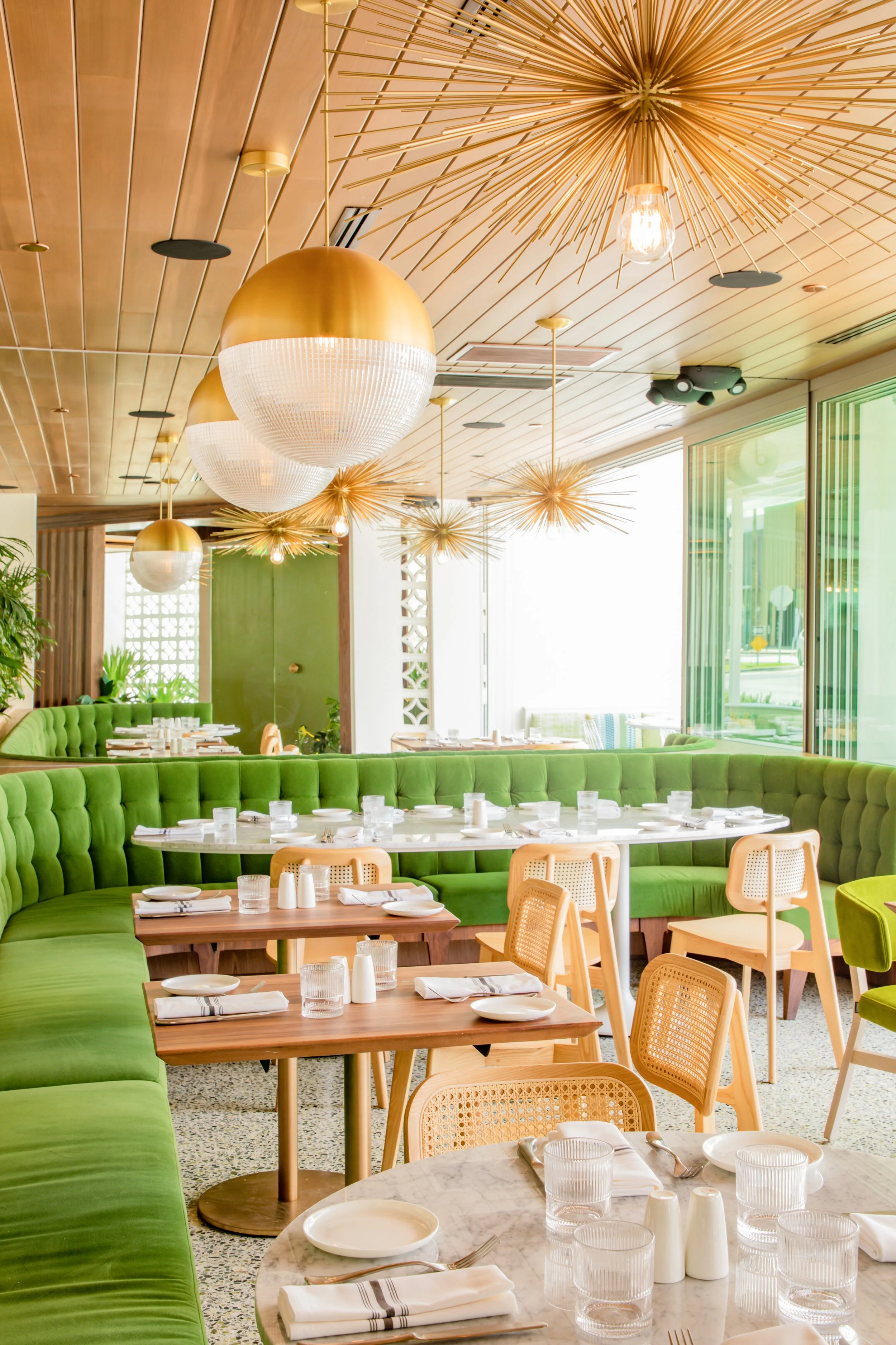 Interior of a bright restaurant with green banquette seating, wooden chairs, and a variety of tables set with plates, glasses, and silverware. The ceiling features modern gold and white pendant lights and starburst chandeliers, and large windows let in natural light.