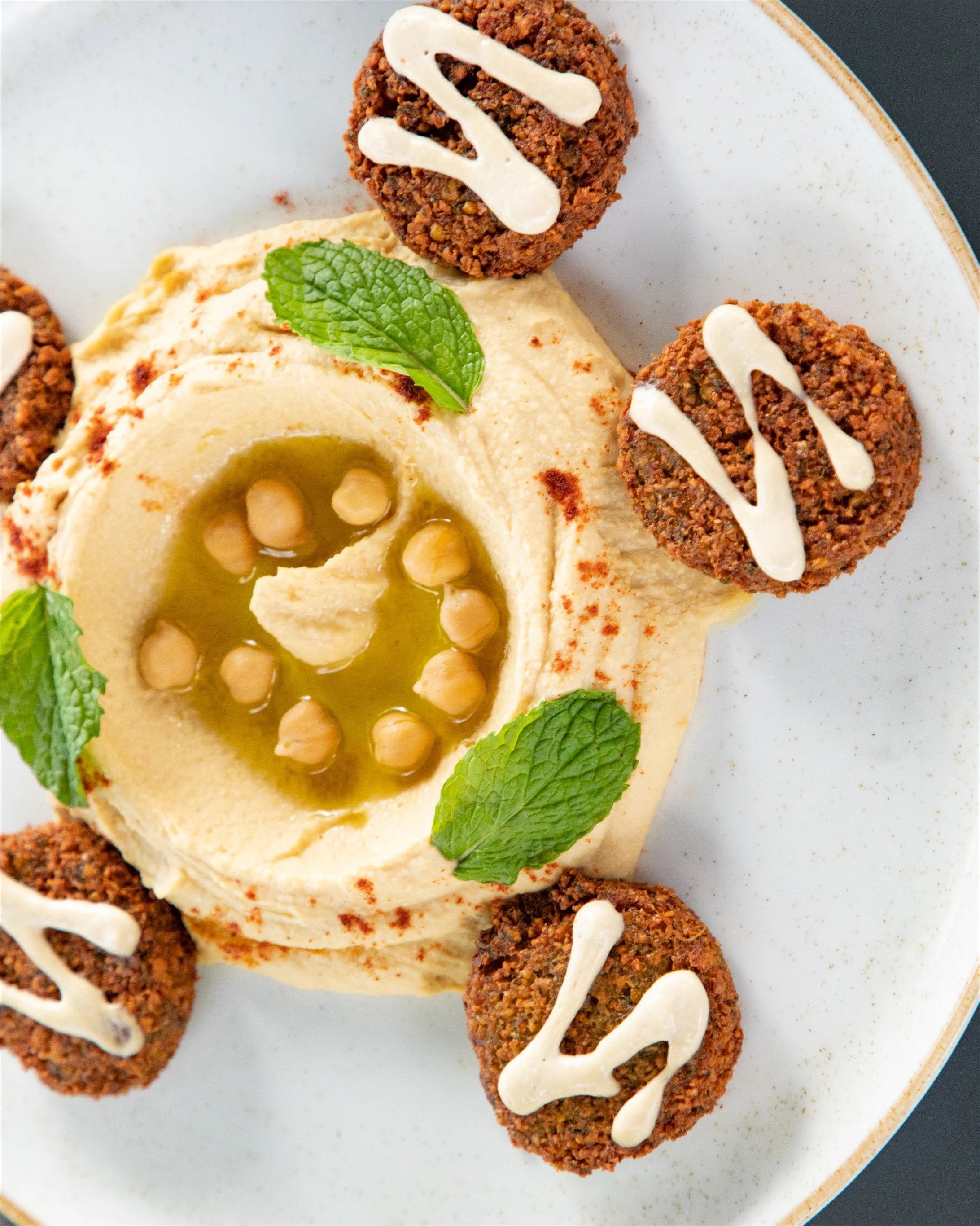Plate with hummus garnished with chickpeas, mint leaves, and paprika, topped with three falafel balls drizzled with white sauce.