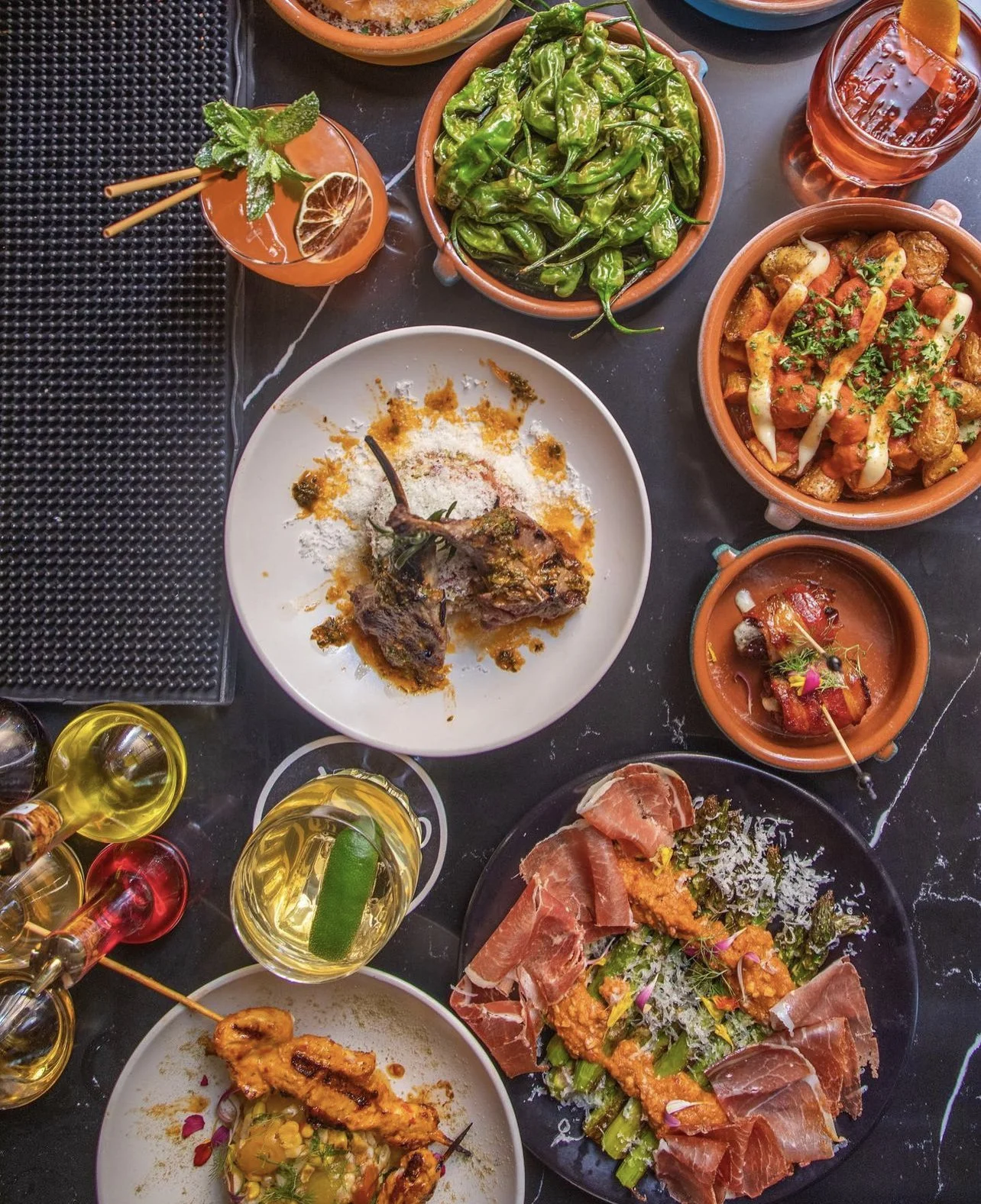 A top-down view of a variety of Mexican dishes on a black table, including a plate with two grilled lamb chops, a bowl of green peppers, a bowl of tinga chicken, a plate with prosciutto and salad, a small bowl of ceviche, and various drinks with lime