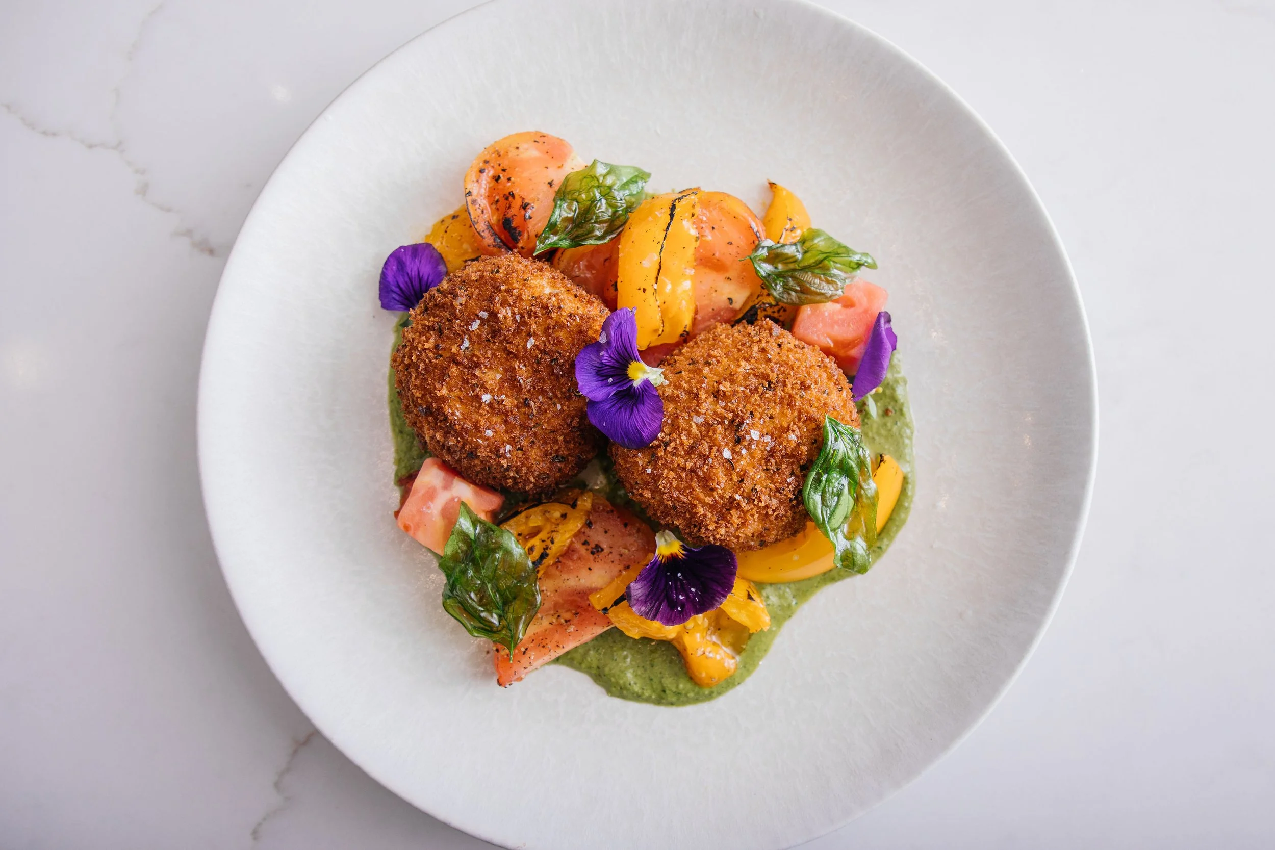 A white plate with two croquettes on a bed of green sauce, garnished with edible purple flowers, and surrounded by chopped tomatoes, yellow bell peppers, and fresh basil leaves.
