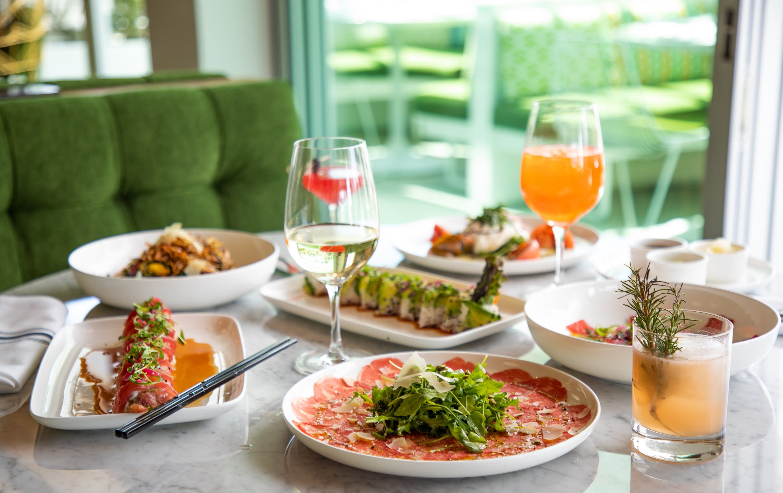 A table set with an assortment of dishes including sushi, sliced meat with greens, a salad, and various beverages in glasses, with a green booth and bright windows in the background.
