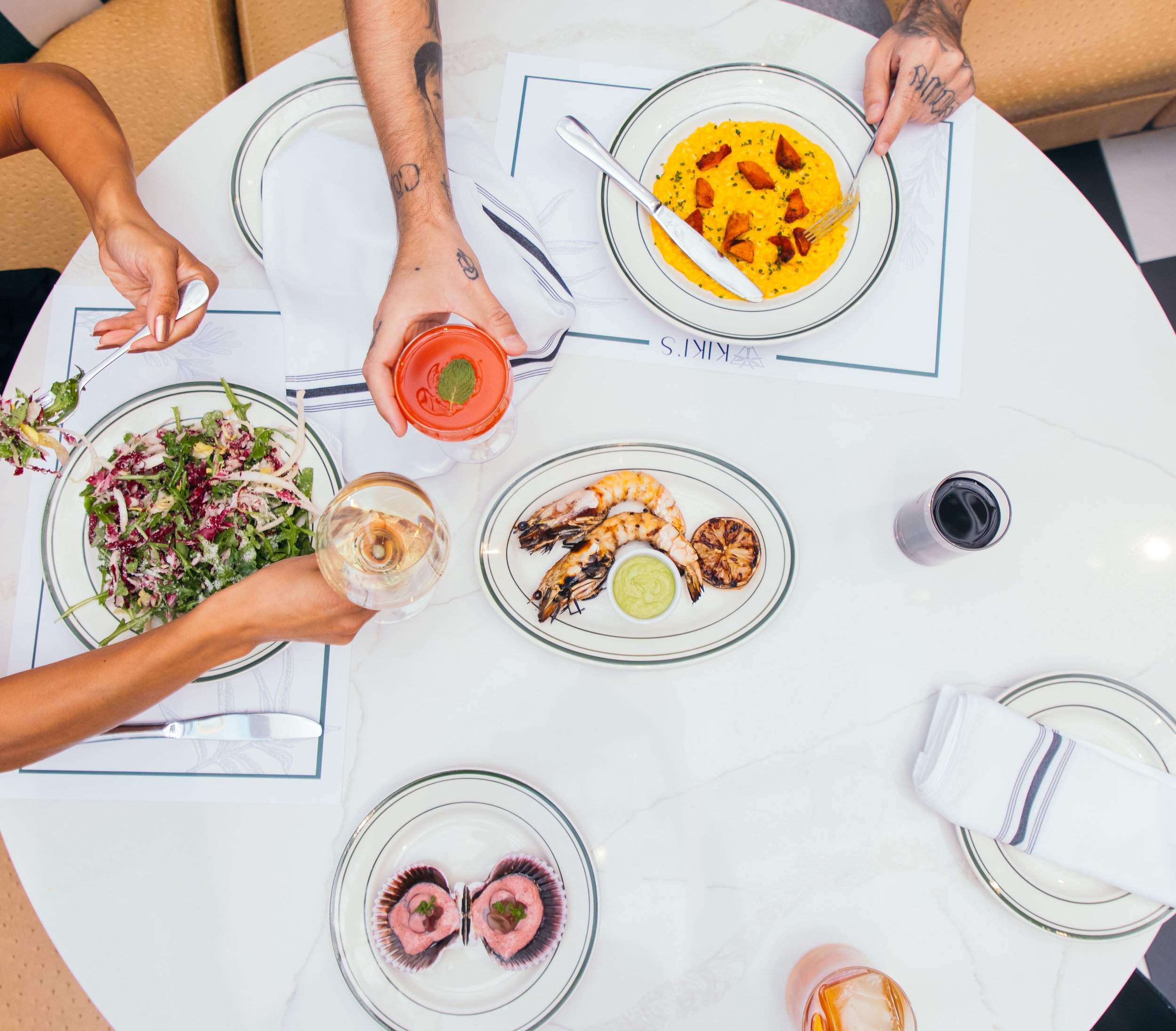 Top-down view of a round dining table with various dishes including a salad, grilled shrimp with lemon, a bowl of yellow curry with chicken, a dessert with pink frosting, and drinks like wine, cocktail, and soda, with hands reaching for food and hold