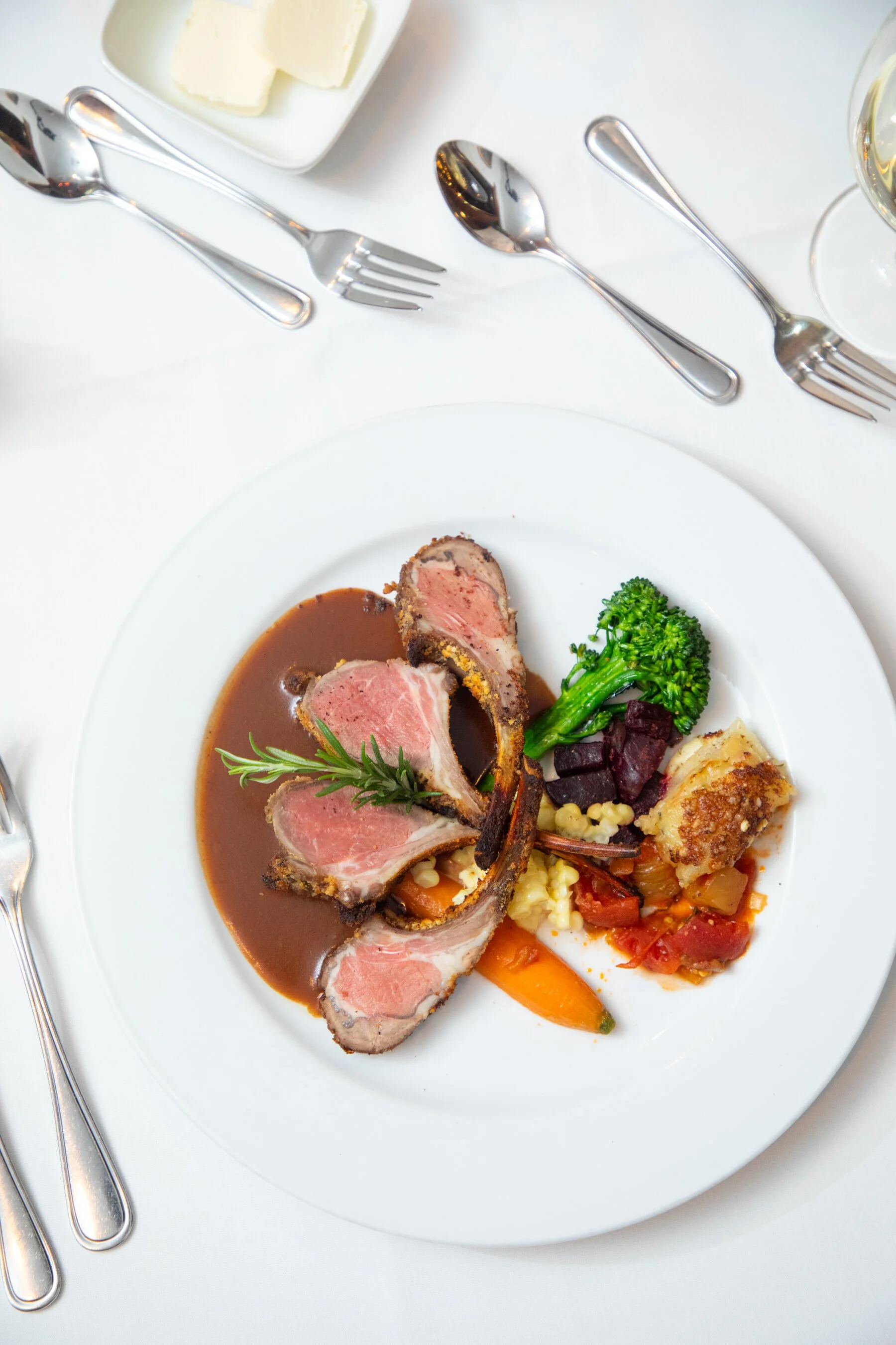 A white dinner plate with sliced rack of lamb served with a brown sauce, accompanied by broccoli, root vegetables, and a potato gratin. The plate is set on a white tablecloth with silverware around it, and a glass of white wine is partially visible.