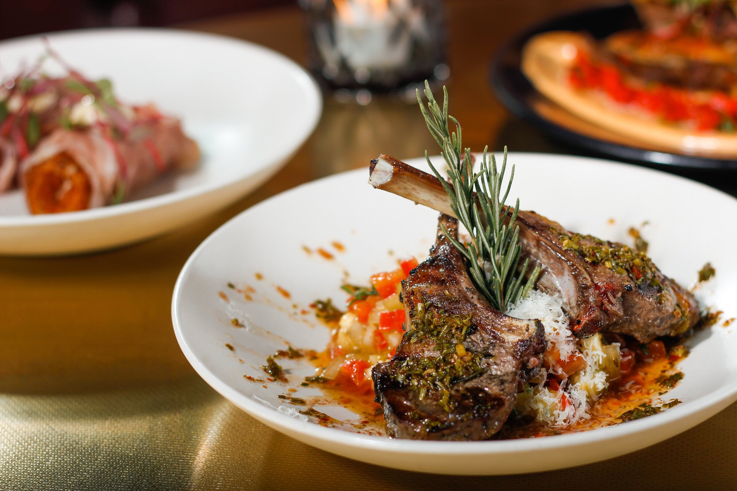A plated lamb chop garnished with rosemary, served over a bed of diced vegetables and sauce.