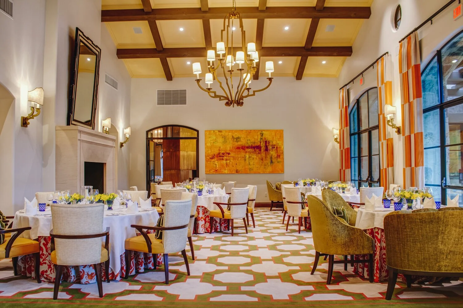 Elegant restaurant dining room with round tables set with white tablecloths, floral centerpieces, and glassware, beige and green armchairs, large windows with orange and white curtains, a chandelier hanging from a wooden beam ceiling, and an abstract