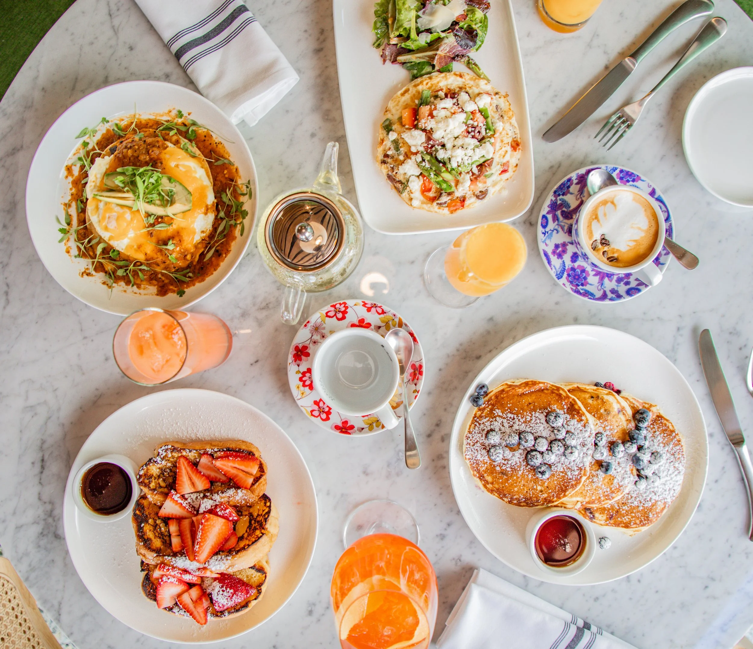 A breakfast spread on a marble table includes pancakes with blueberries, strawberries, and powdered sugar, a fried egg-topped dish with herbs, a pizza with vegetables and cheese, a salad, various drinks, and utensils.