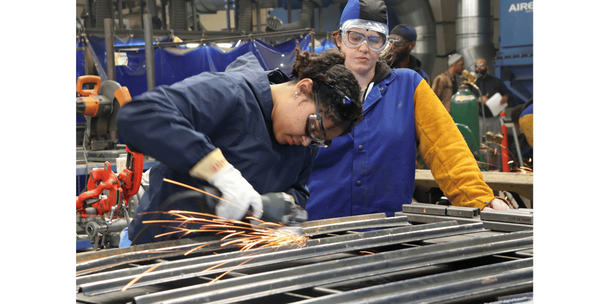 An image of two women at a trades or CTE school. An apprentice and her mentor grinding and welding.
