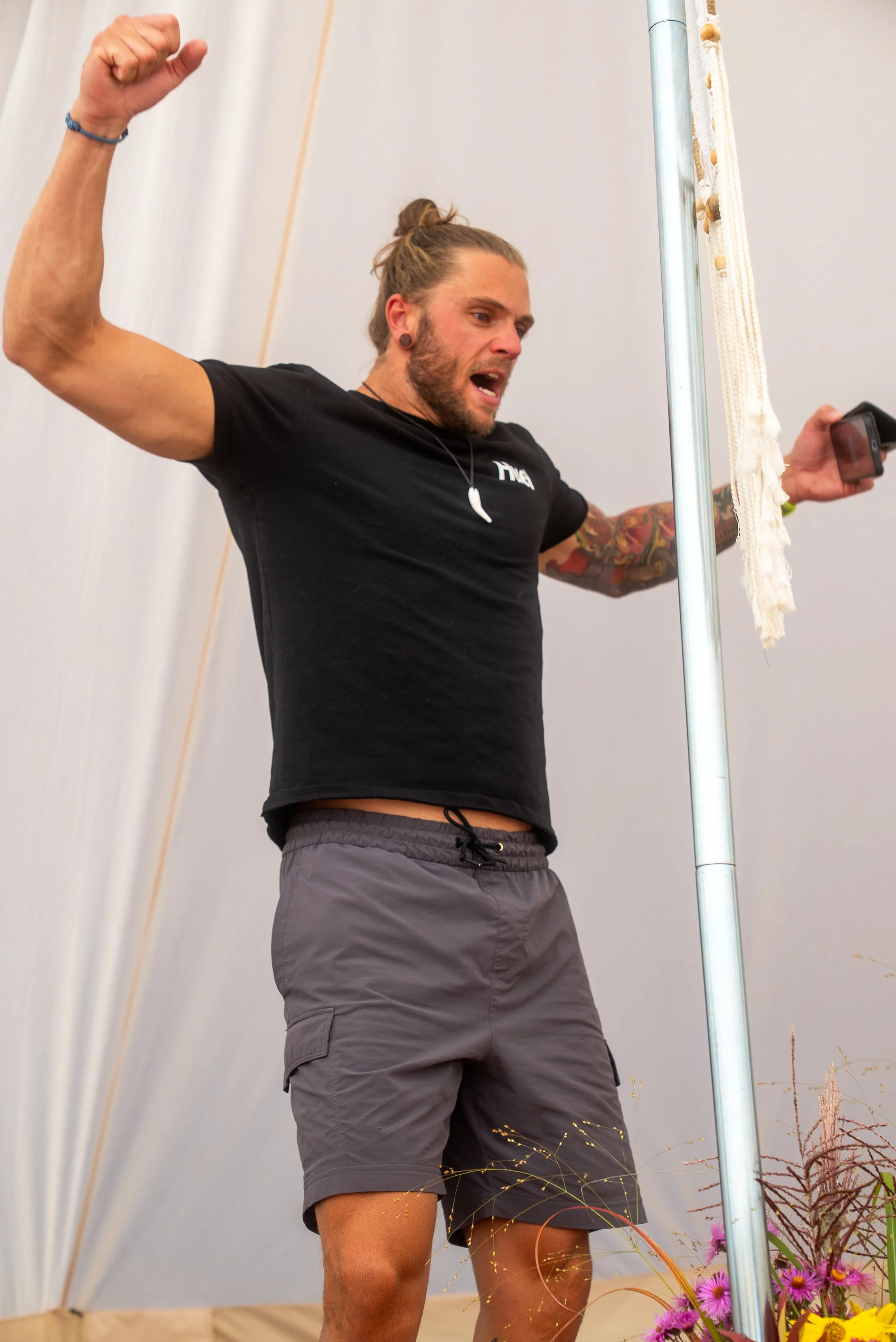 A man with long hair in a bun, wearing a black t-shirt, gray shorts, and a bracelet, raising his fist in the air while holding a phone in his other hand, standing indoors near a metal pole with decorative hanging beads and a floral arrangement at his feet.