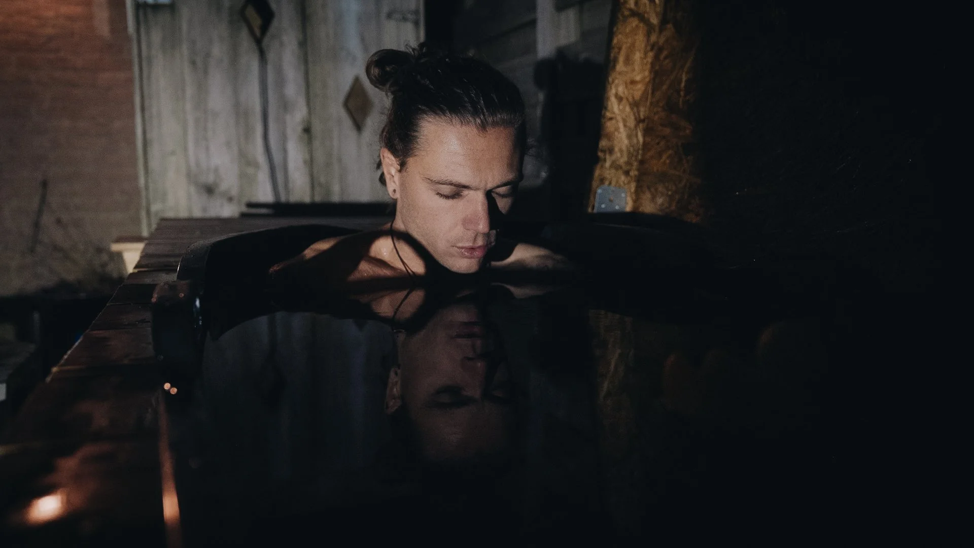 A man with long hair in a bun is resting his head on a piano, with his eyes closed, in a dimly lit room with wooden walls.