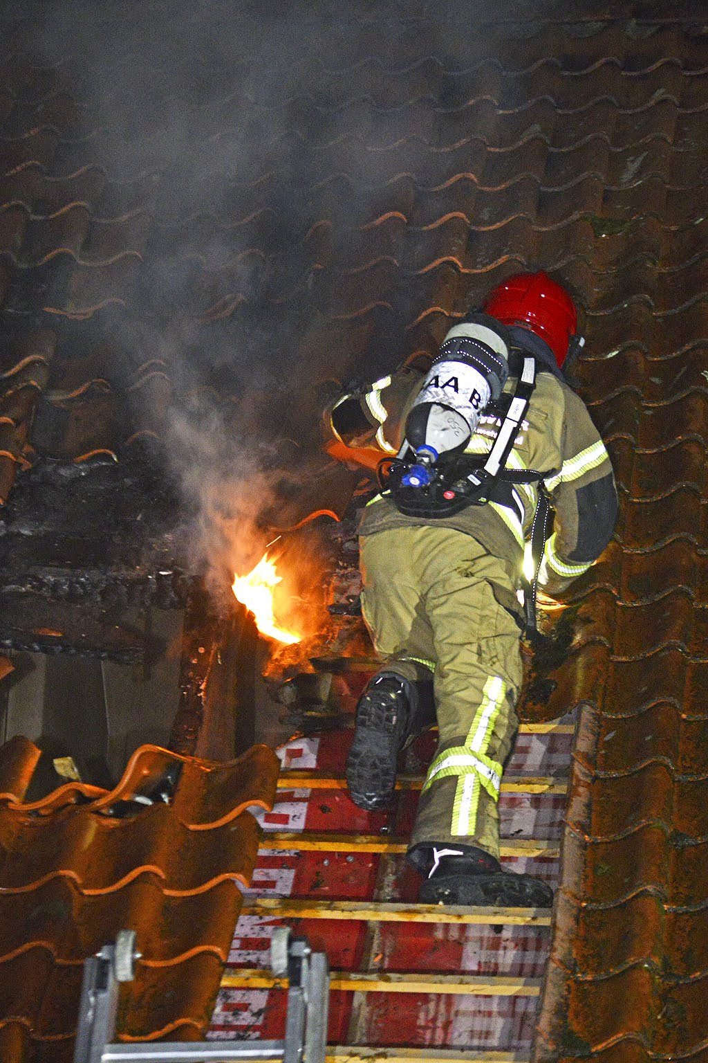 Firefighter in full gear fighting a fire on a building's roof at night, with flames and smoke visible.
