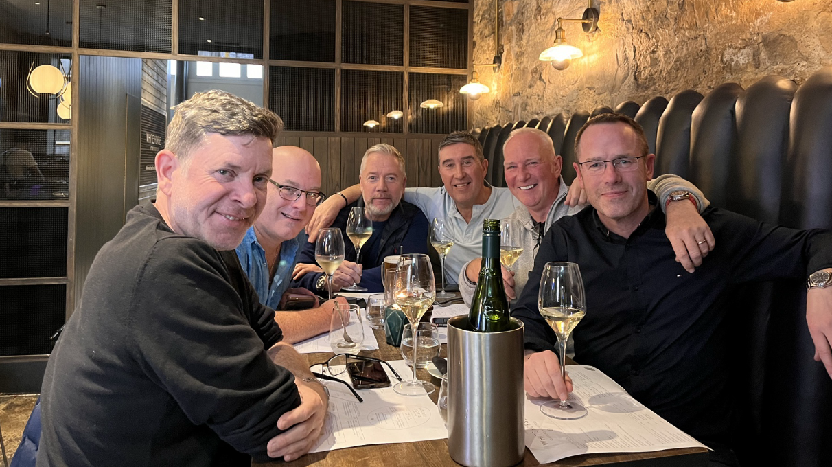 Six men sitting around a dining table in a restaurant, smiling and holding wine glasses, with menus and a bottle of wine on the table.