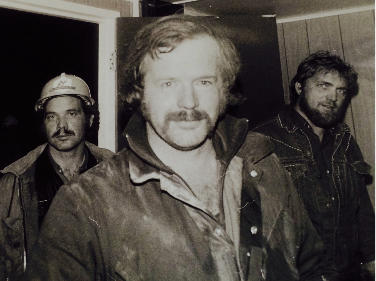 Three men with rugged appearance, one wearing a helmet, in what appears to be a dimly lit indoor setting.