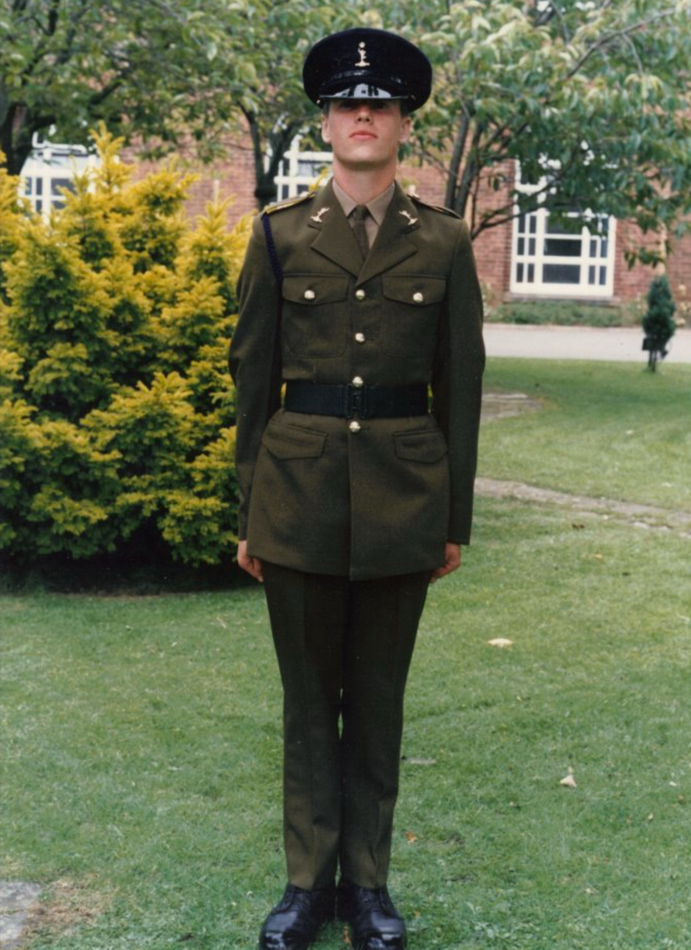 A person in a military uniform standing outdoors on a grassy area, with bushes and trees in the background.