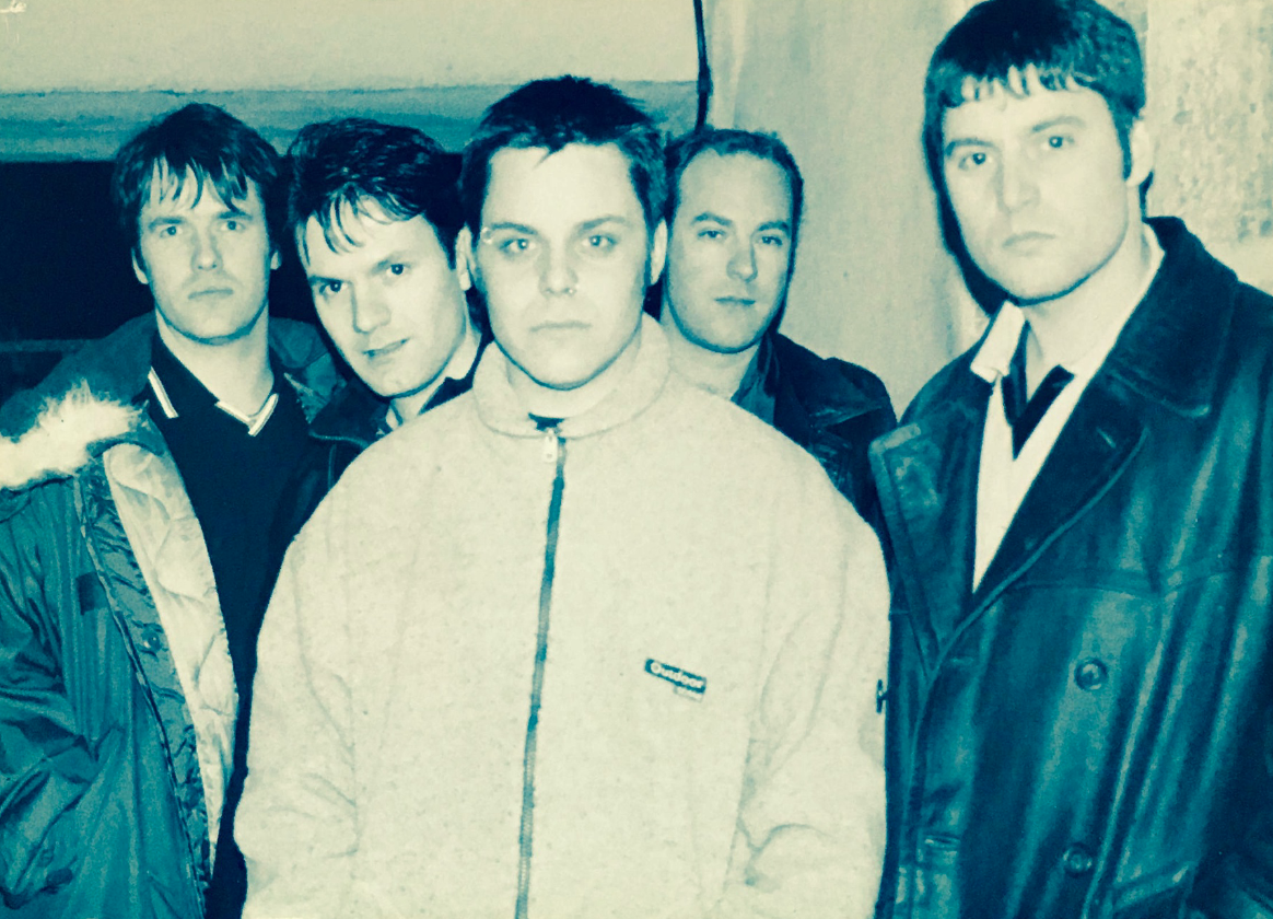 Black and white photo of five young men standing close together, looking directly at the camera. They are wearing jackets and sweaters, and are positioned in a casual indoor setting.