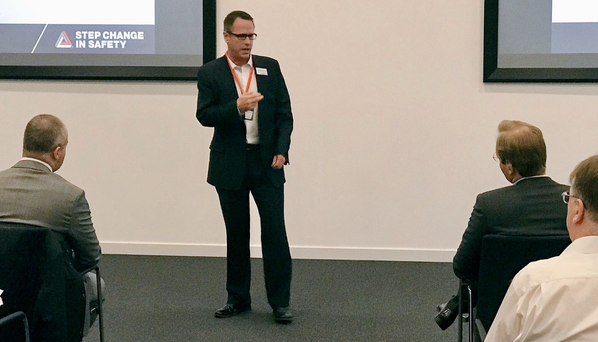 Man in glasses and dark suit giving a presentation to an audience in a conference room, with a slide titled 'Step Change in Safety' projected behind him.