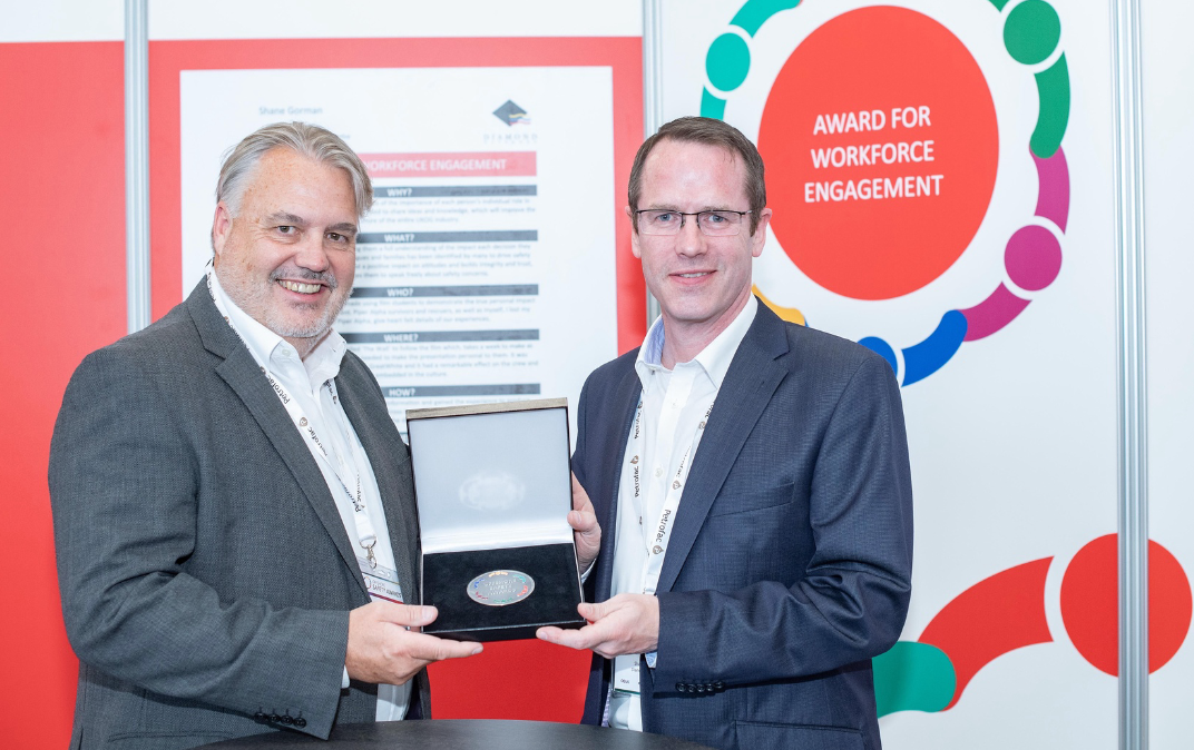 Two men in suits standing at a conference booth with a red poster that says "Award for Workforce Engagement." One man is presenting an award to the other inside an open box.