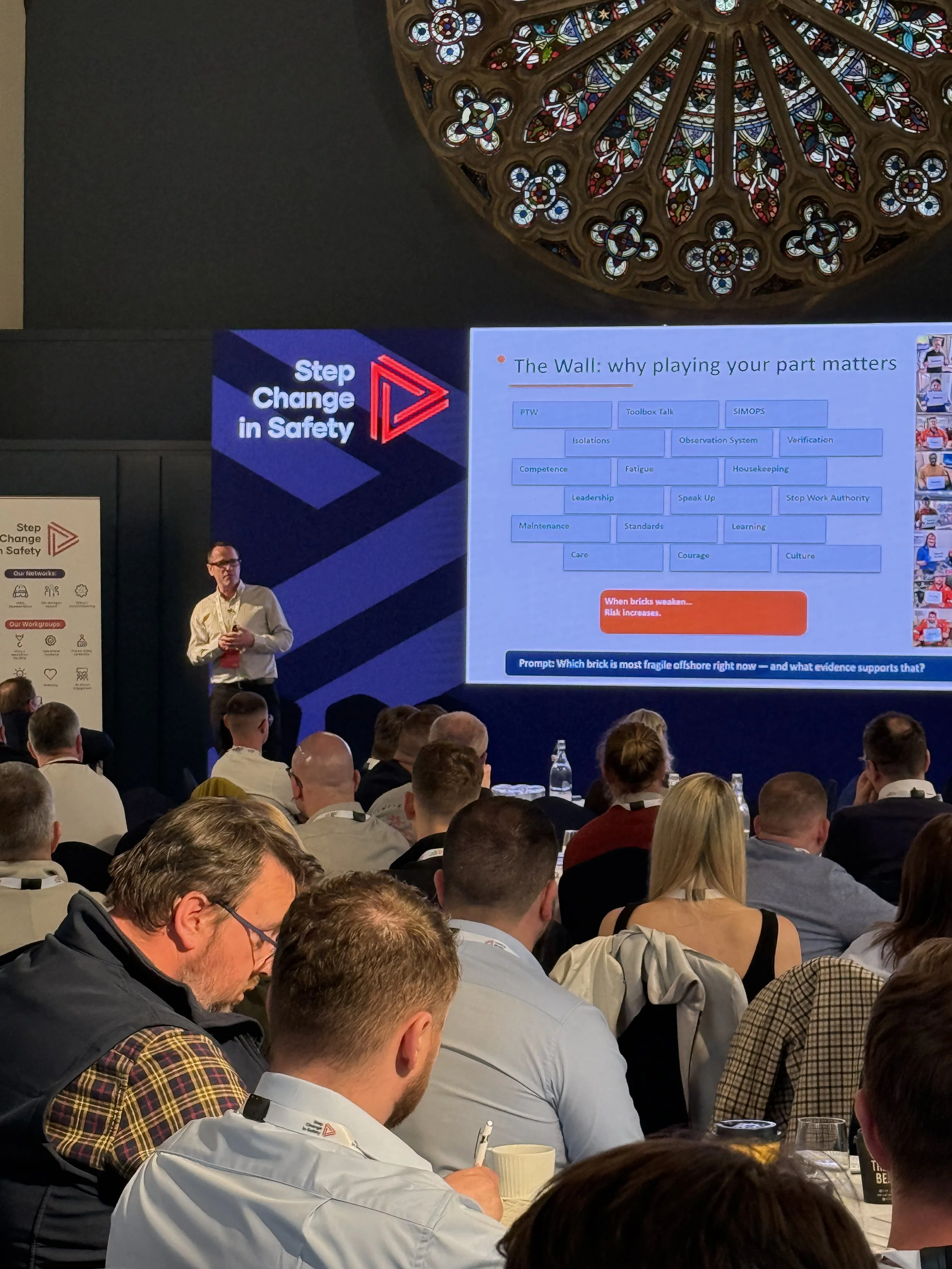 A man in glasses and a white shirt standing and speaking in front of an audience, with a large presentation slide behind him displaying the title 'The Wall: why playing your part matters' and various related keywords, at a conference or seminar. The 