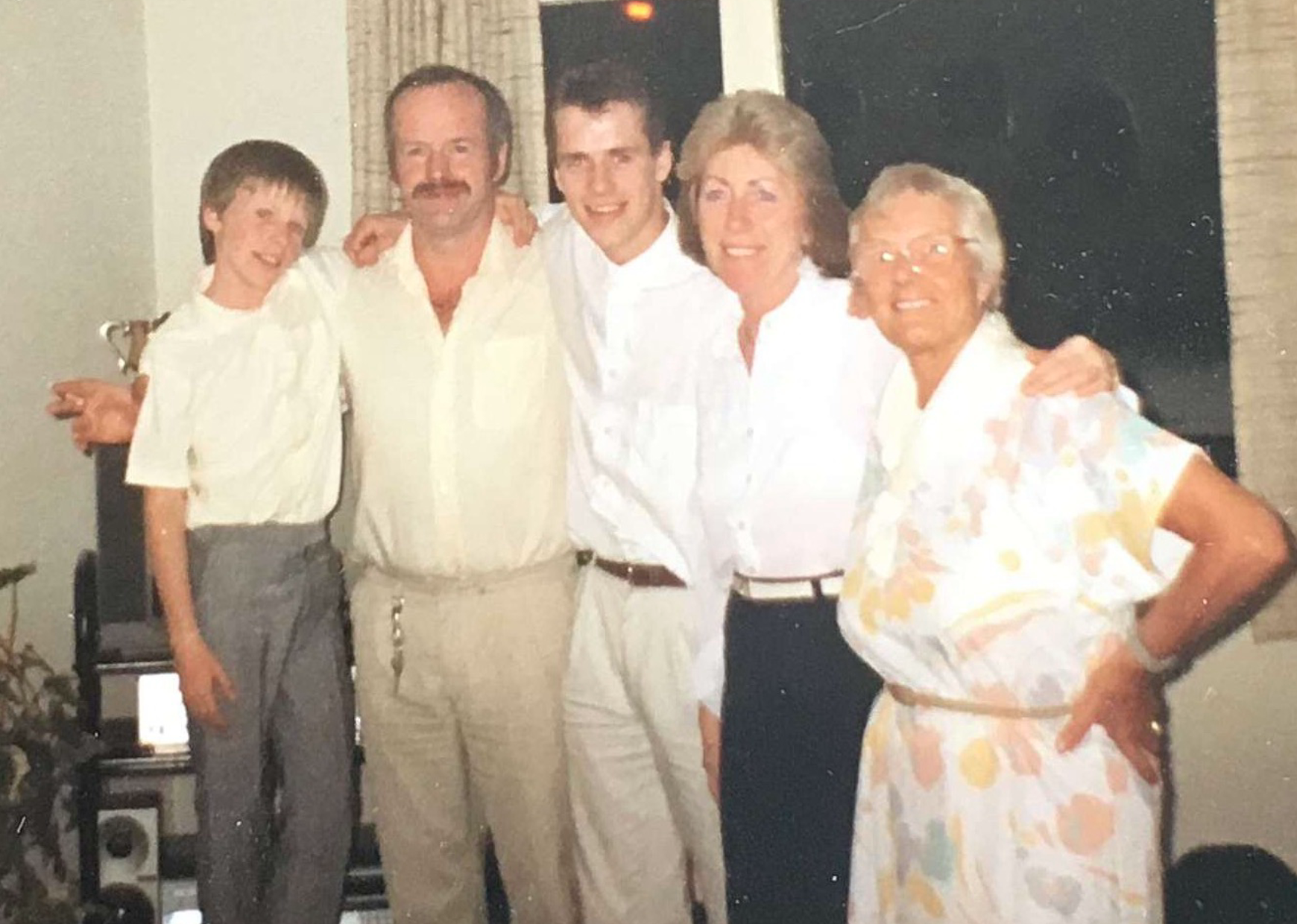 Five people standing indoors, smiling and posing for a photo. They appear to be family members or friends. The group includes two men and three women, all dressed in semi-formal or casual clothing. The background shows a window with curtains.