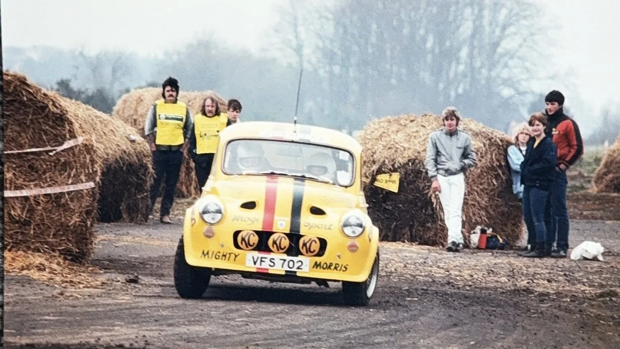 A small yellow rally car with a red and black striped design on the hood, surrounded by hay bales, with five people standing in the background.
