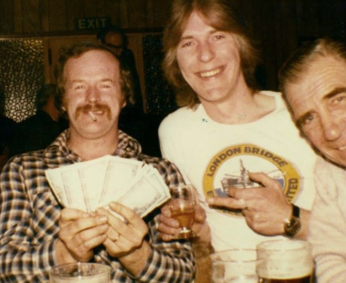 Three men at a bar, with one holding several dollar bills, another pointing at his T-shirt with a London Bridge logo, and the third smiling. The man on the left has a mustache, is holding money, and has a cigarette in his mouth. The man in the center