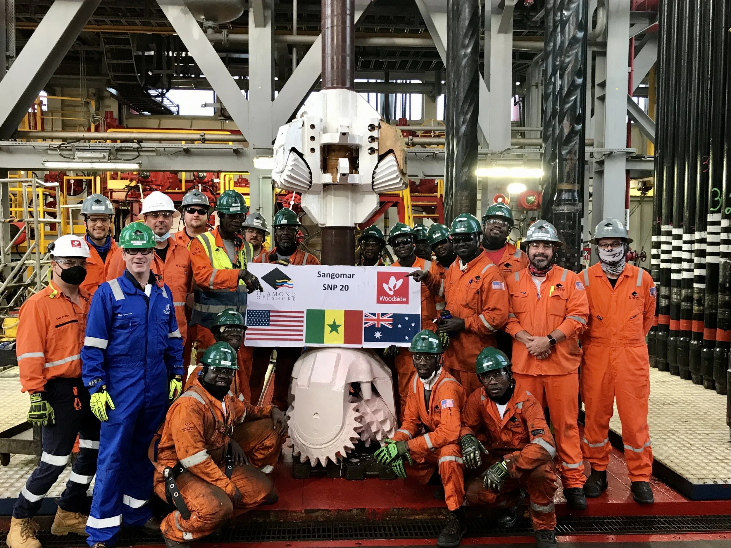 Group of workers in orange safety uniforms and hard hats holding a banner that reads 'Sangomar SNP 20' in an industrial facility with machinery and pipes in the background.
