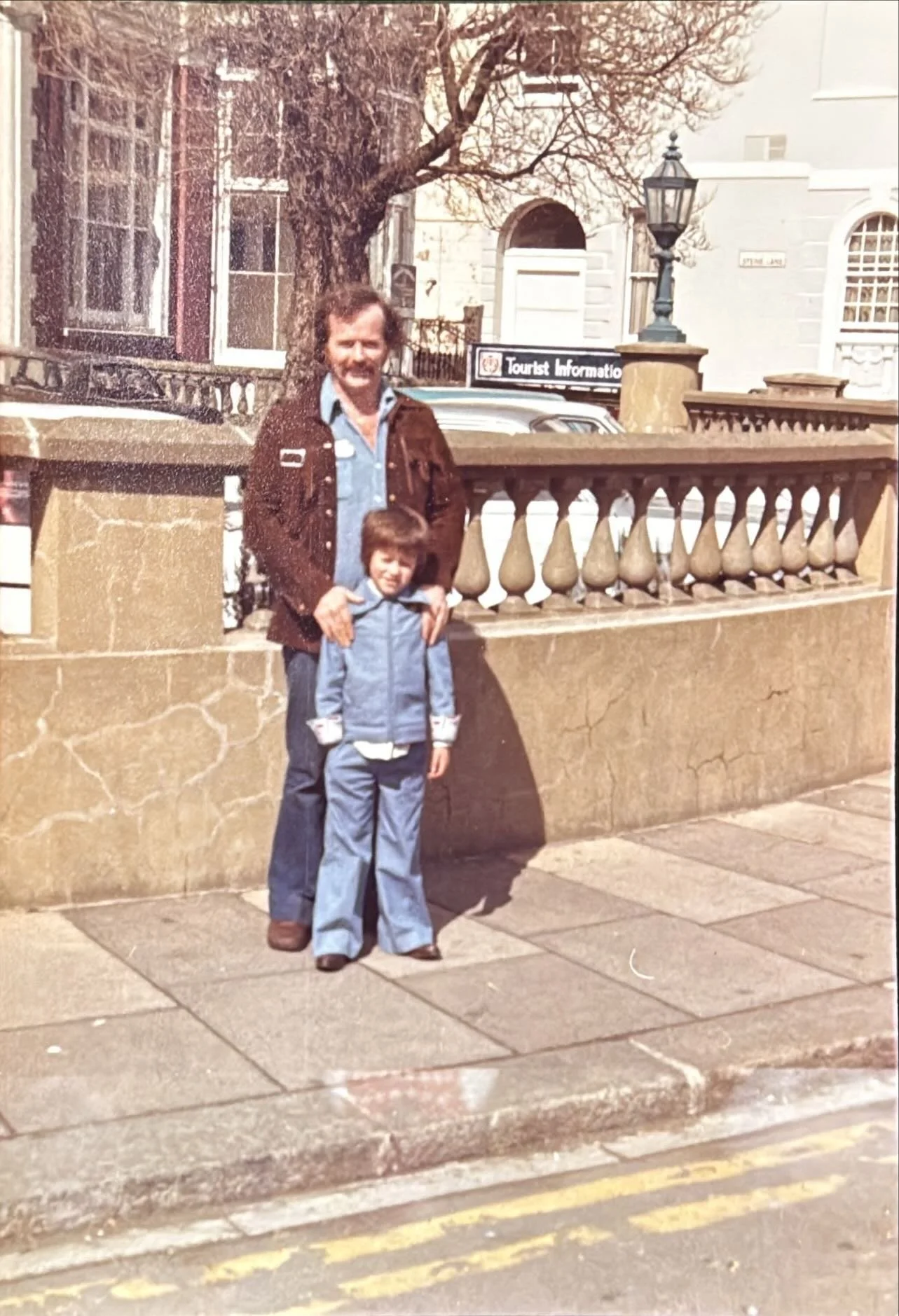 A man and a young boy standing outside near a stone wall and a sidewalk, with historical buildings and a street lamp in the background. The man is wearing a brown jacket and the boy is dressed in a light blue jacket and pants.