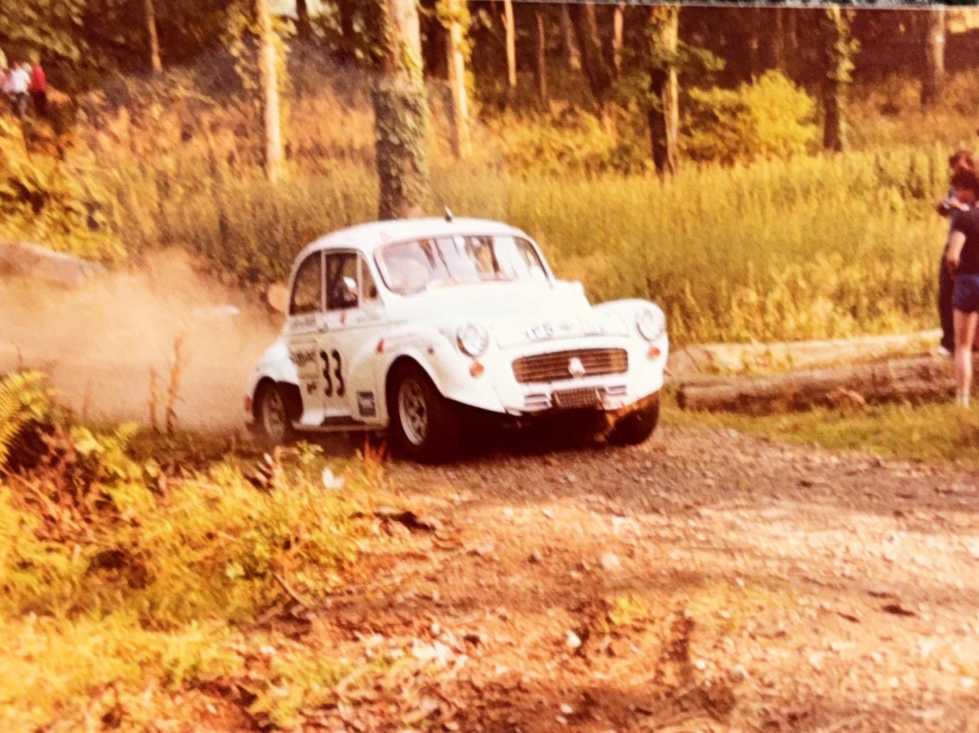 A vintage white rally car with the number 33 racing on a dirt trail through a forested area with yellow autumn leaves, kicking up dust behind it. There are people on the side of the trail watching the race.