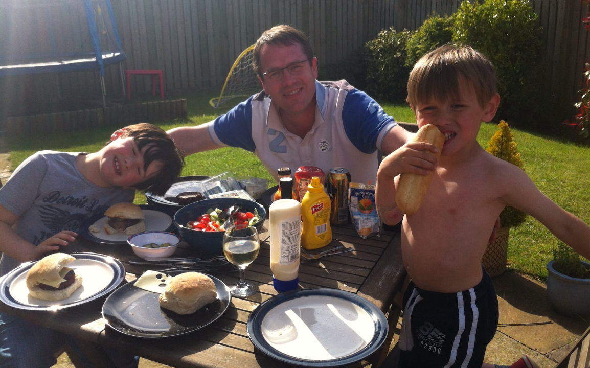 A family enjoying a backyard barbecue with kids eating hot dogs and beef sandwiches at a picnic table filled with food and drinks.