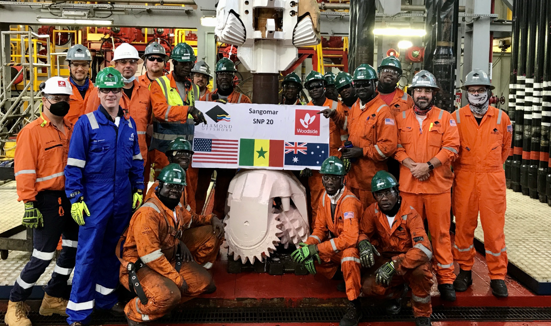 Group of workers in safety gear holding a sign labeled 'Sangomar SNP 20' with flags of the United States, Senegal, United Kingdom, Australia, and a logo for Woodside, inside an industrial oil or gas facility.