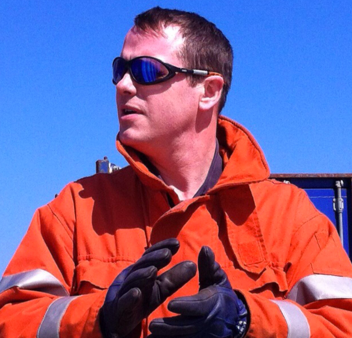 A man wearing orange safety gear, black gloves, and sunglasses outdoors against a blue sky.