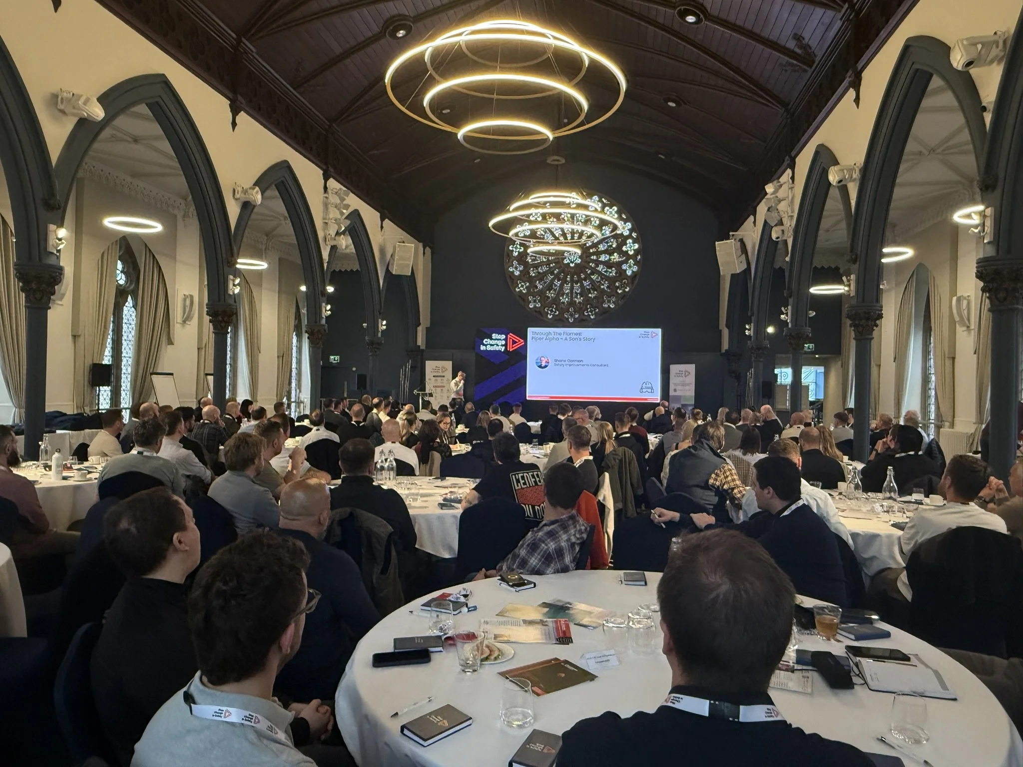 A large indoor conference hall filled with attendees sitting at round tables. The front of the hall has a stage with a speaker giving a presentation. The room features ornate architecture with high vaulted ceilings, decorative columns, and large stai