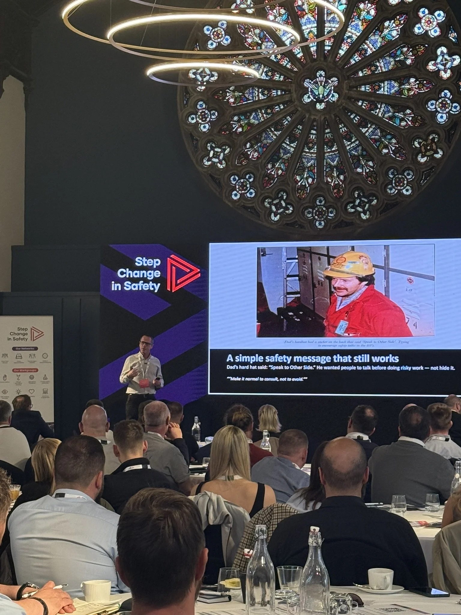 A speaker in a white shirt and glasses is giving a presentation at a conference with a large audience. The presentation slide on the screen contains a picture of a worker wearing a yellow hard hat and red jacket, with a safety message about speaking 