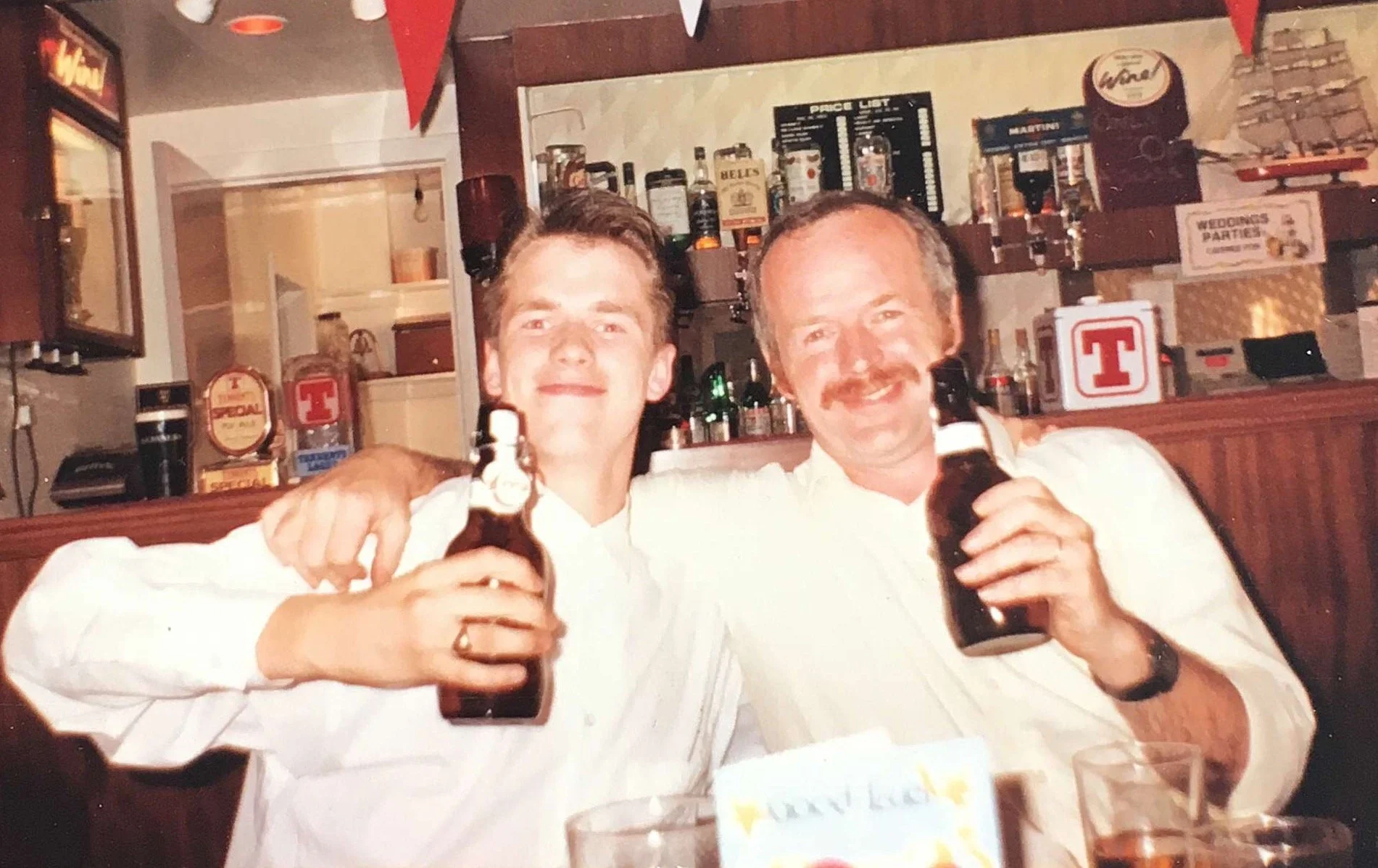 Two men sitting at a bar, smiling, holding beer bottles. They are wearing white shirts. The bar has various bottles and glasses behind them, with a sign reading 'WEDINGS PARTIES'.