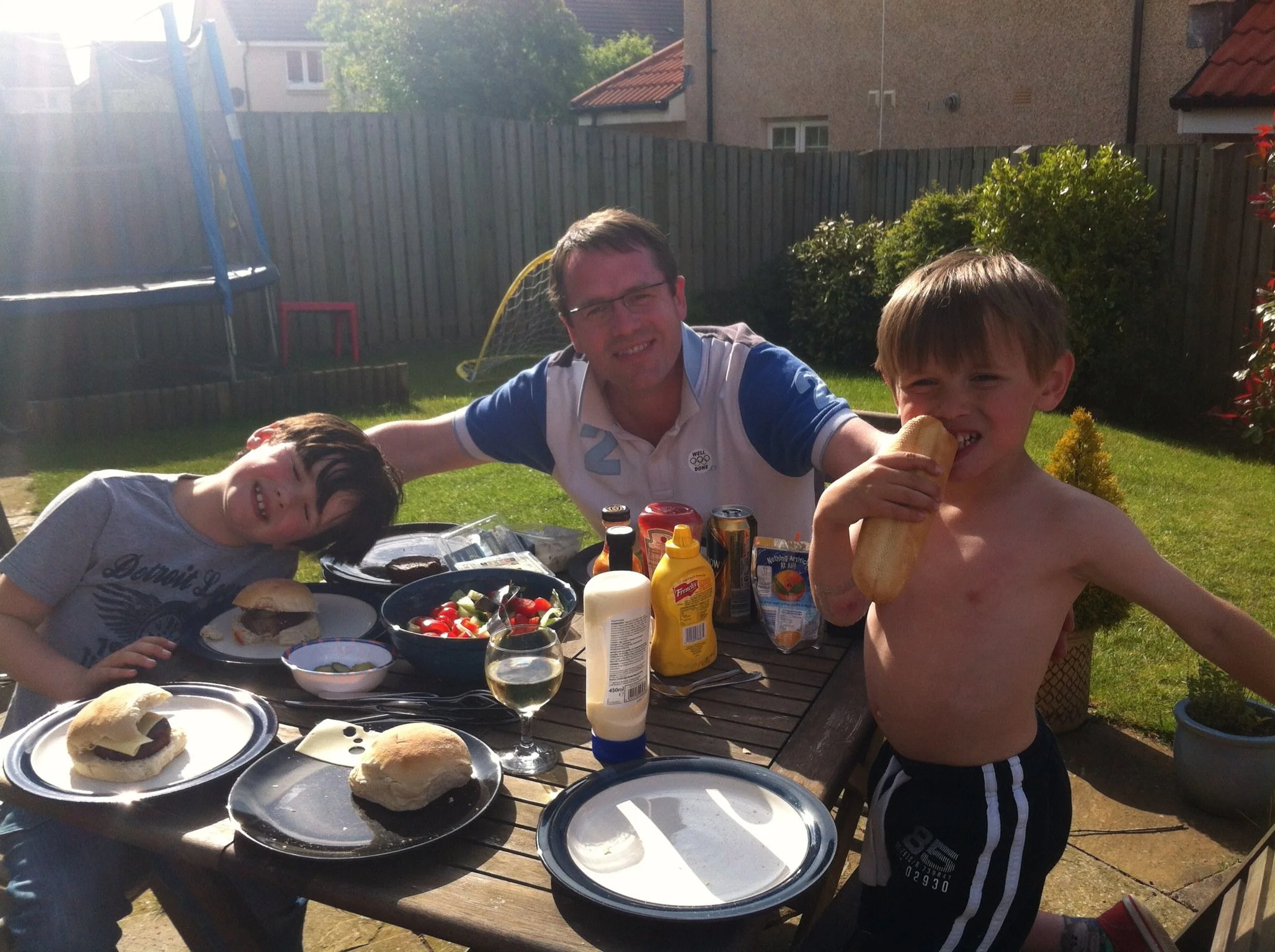 Family having a barbecue in a backyard on a sunny day, with plates of burgers, salad, and condiments on the table.