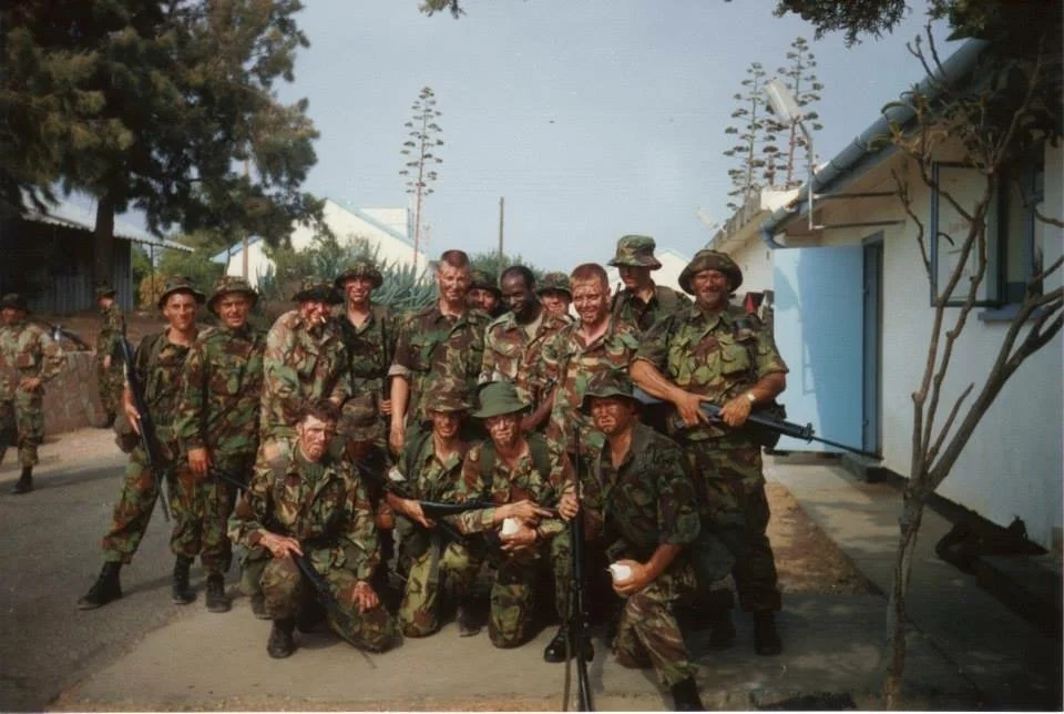 Group of soldiers in camouflage uniforms posing outdoors, some holding rifles, near a white building with trees in the background.